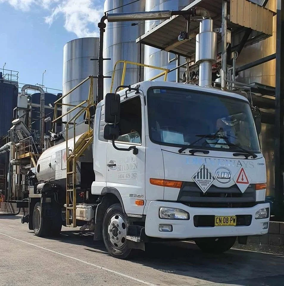 A white utility truck parked on the street, with warning signs on the front and a yellow safety ladder on the side, and industrial tanks and piping in the background.