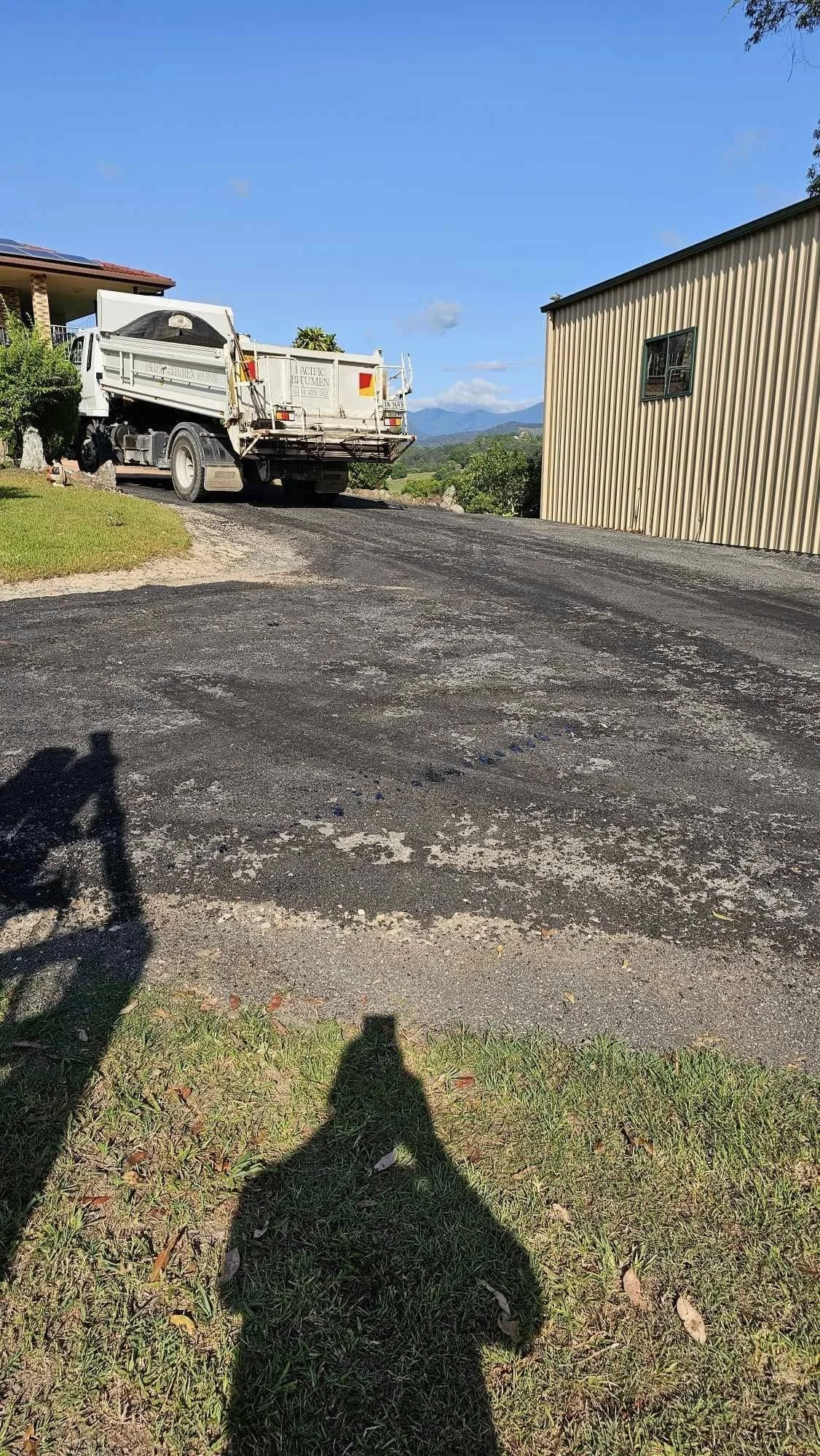A truck parked on a newly paved driveway next to a house with a tin wall, and a green lawn in the foreground. Mountain scenery in the background under a clear blue sky.