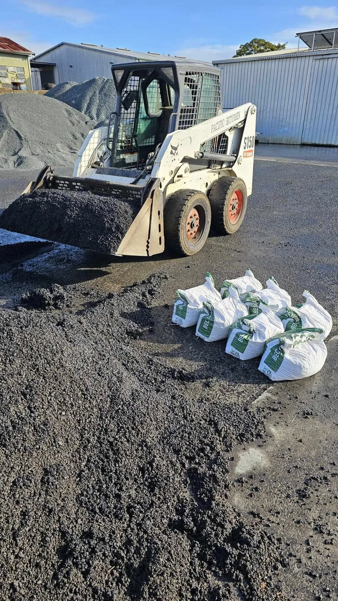 A skid-steer loader on a construction site with piles of gravel and sand, and several white bags of building material on the ground.