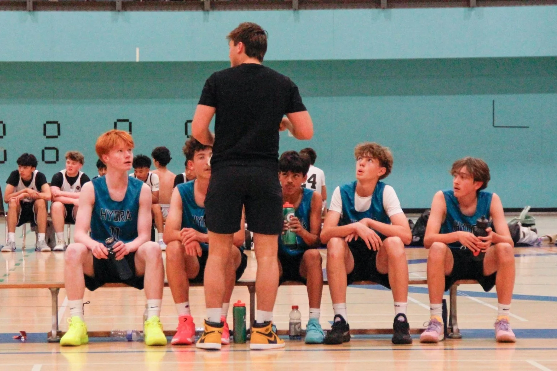 A group of young male basketball players sitting on a bench during a game or practice, listening to a coach who is standing and giving instructions. The players are wearing blue uniforms and some are holding water bottles.