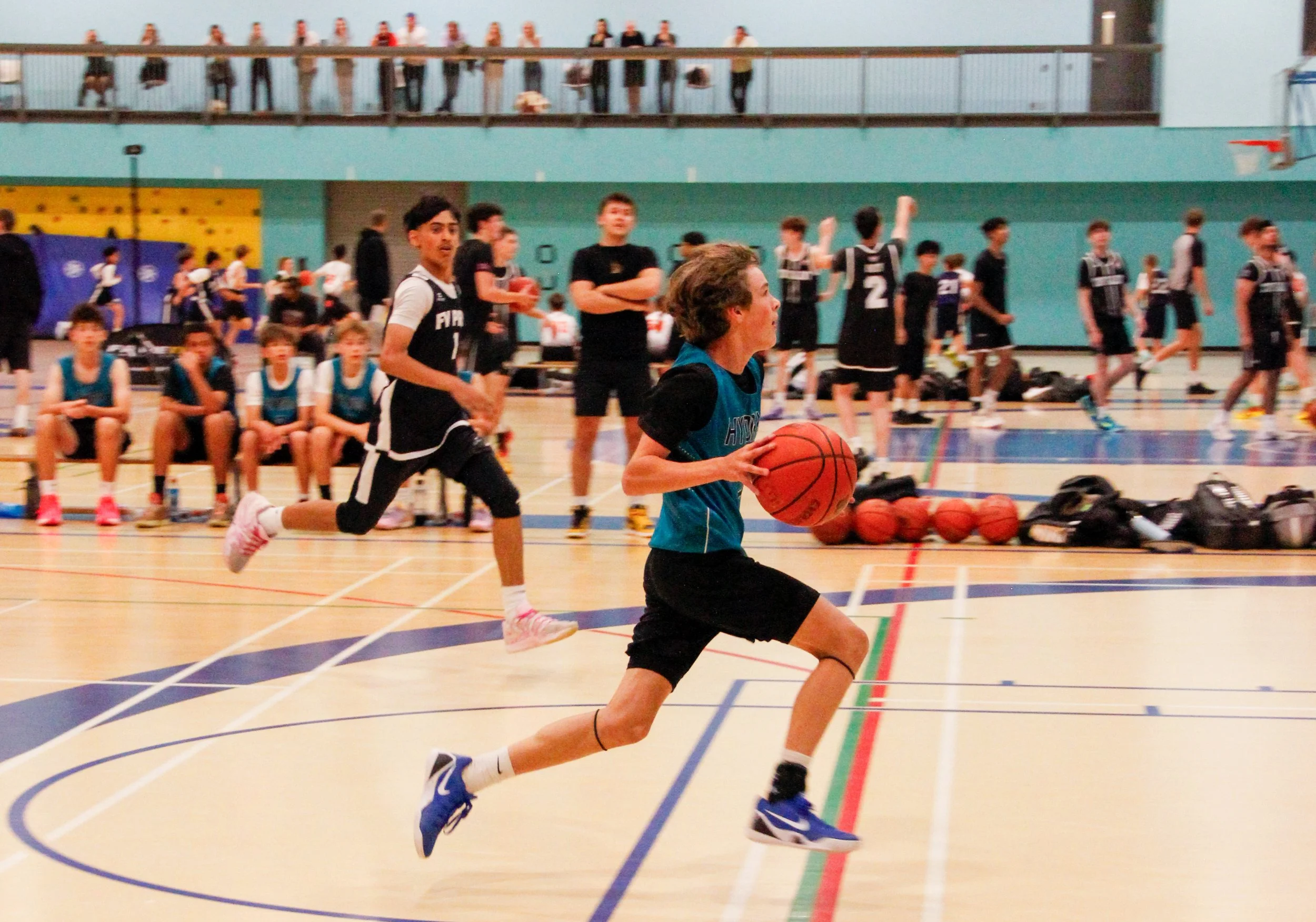 Young basketball player dribbling the ball on an indoor court during practice or game, with other players and spectators in the background.