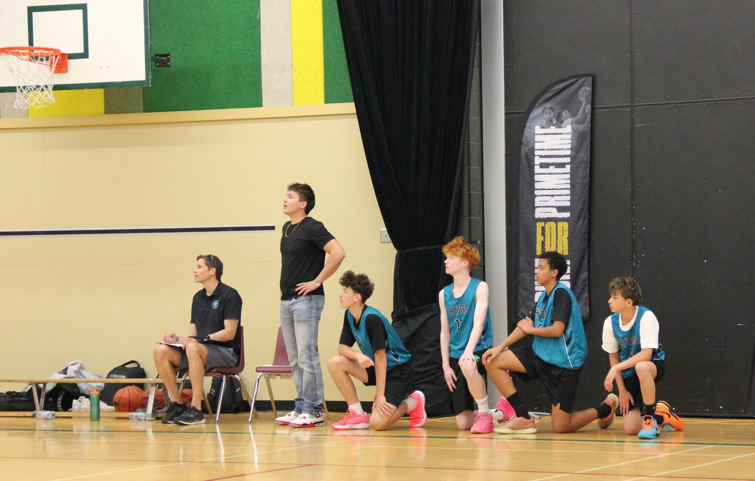 A youth basketball team during a game or practice session, with some players kneeling on the floor and others sitting on chairs, in an indoor gymnasium. An adult coach is sitting, and a young standing player appears to be giving instructions or speaking. The players are wearing blue uniforms, and there are basketballs and sports bags on the floor nearby. The gym has padded walls and a banner that reads 'PRIDE FOR' on one side.