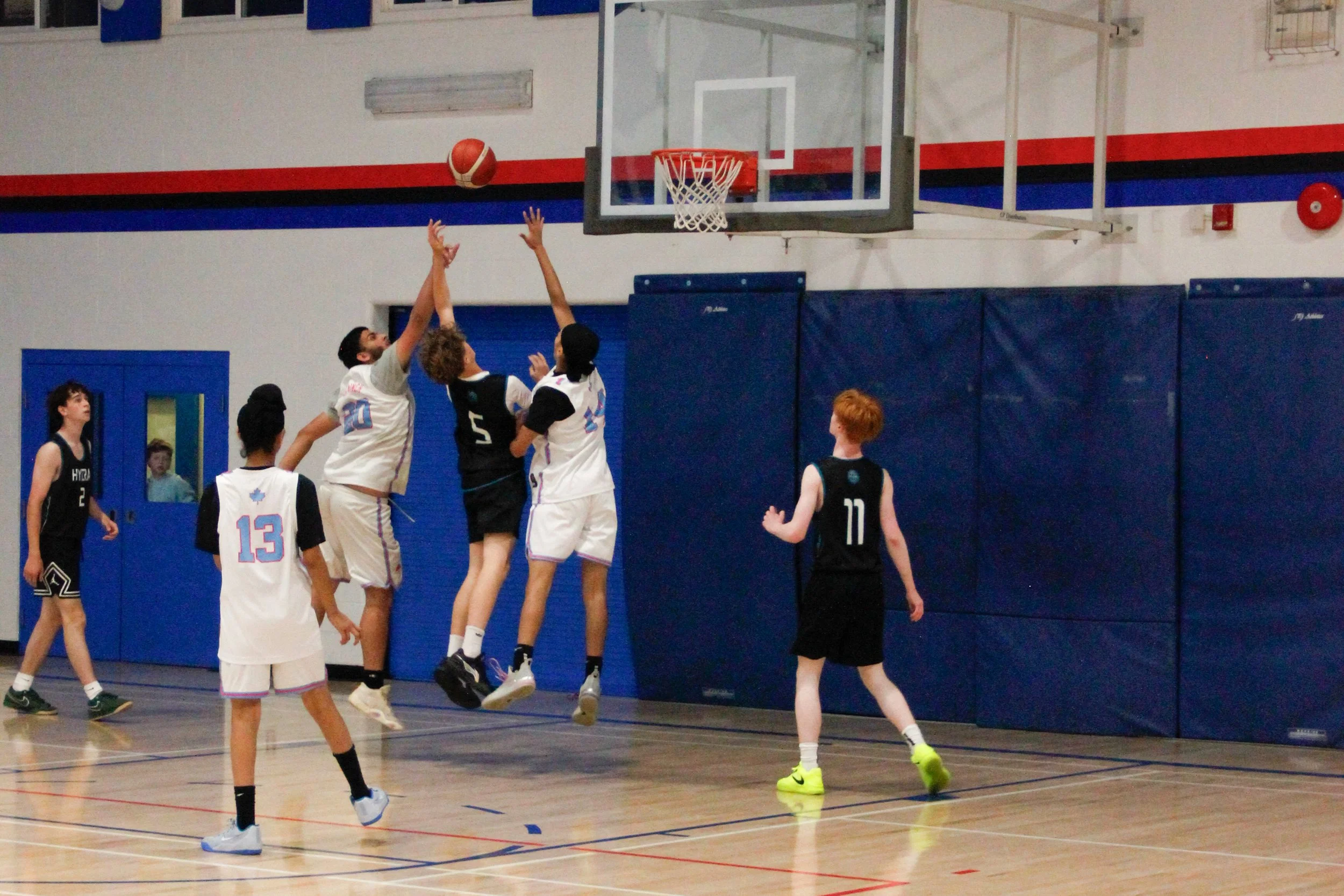 Young basketball players in action during a game inside a gymnasium. One player is jumping to make a shot while others are watching and preparing to rebound or defend.