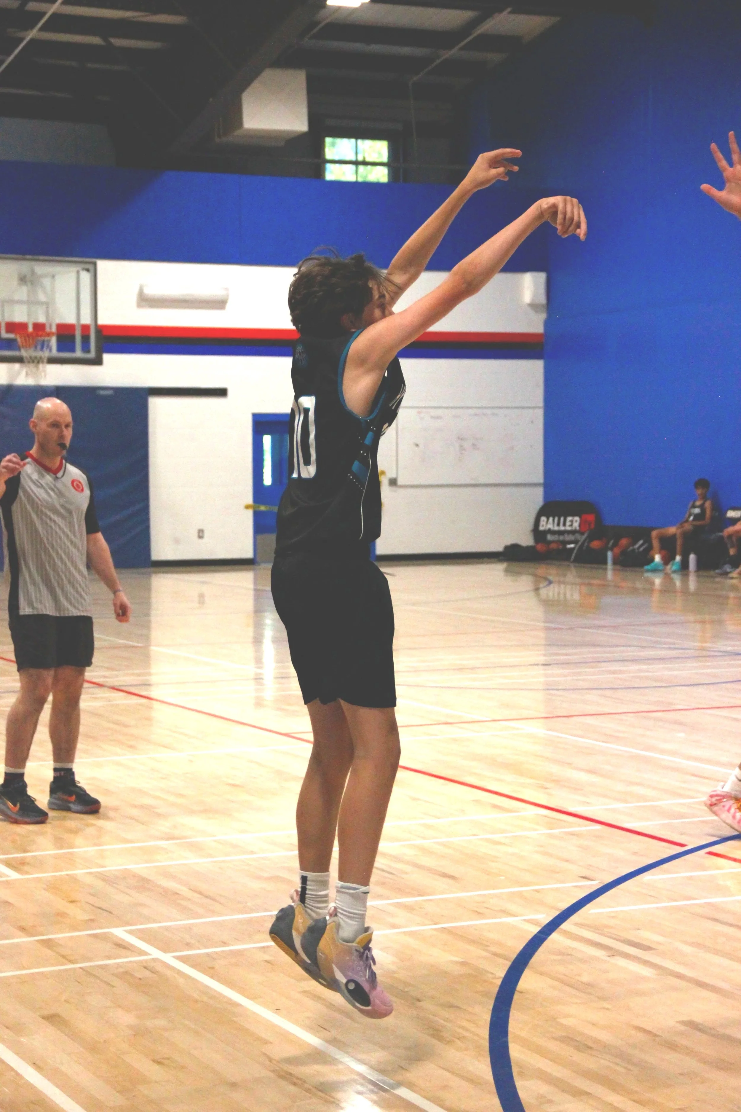 A young man jumping and shooting a basketball in an indoor gymnasium, with a referee and basketball hoop visible in the background.