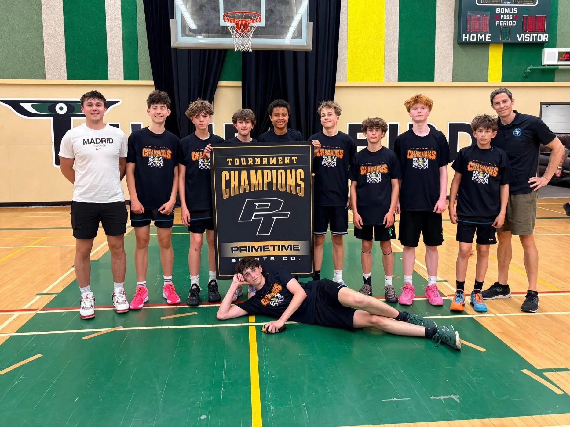 A group of young boys and one adult man standing in a gymnasium, holding a sign that says 'Tournament Champions'. The boys are wearing black T-shirts with a basketball net and ball graphic. There is a basketball hoop and a scoreboard in the background.