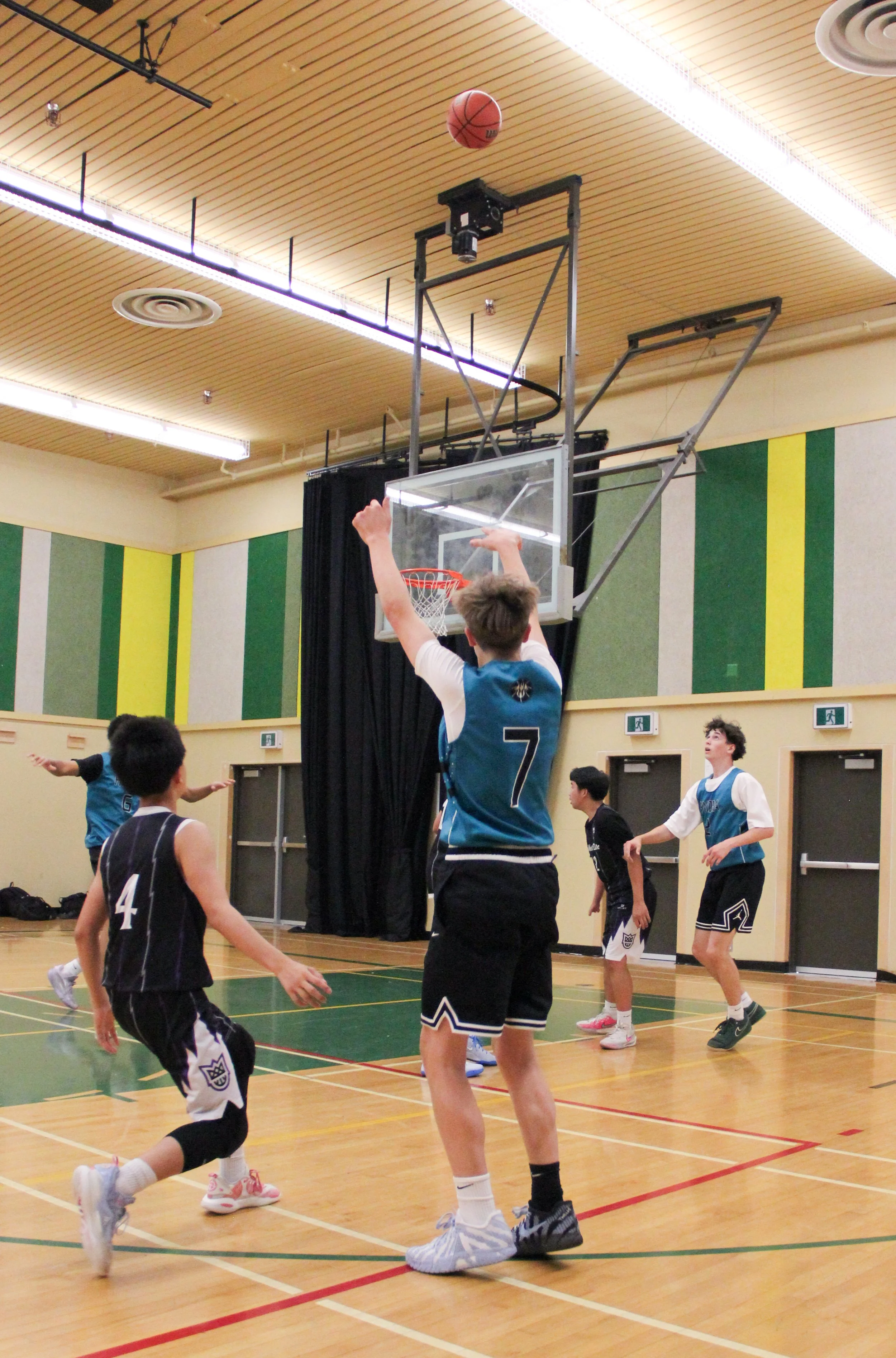 Young boys playing basketball indoors on a court with a yellow and green wall, some boys wearing black, white, and blue jerseys, and a basketball bouncing in the air.