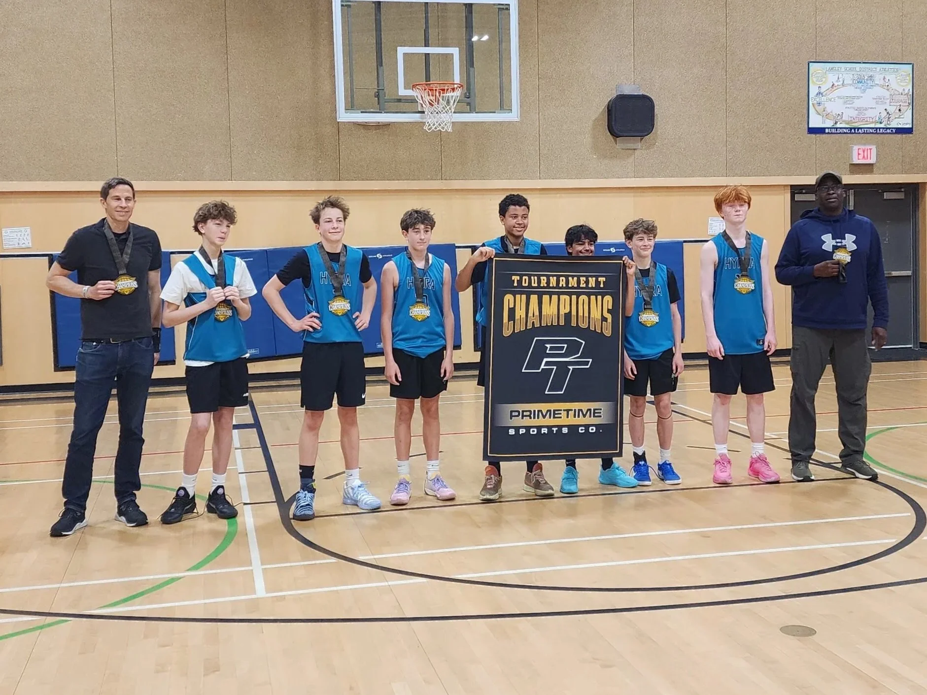 A group of young boys and two adult coaches or referees standing in a gymnasium after a basketball game, holding medals and a championship banner that reads "Tournament Champions." The boys are wearing blue basketball jerseys, and the gym has a basketball backboard and scoreboard.