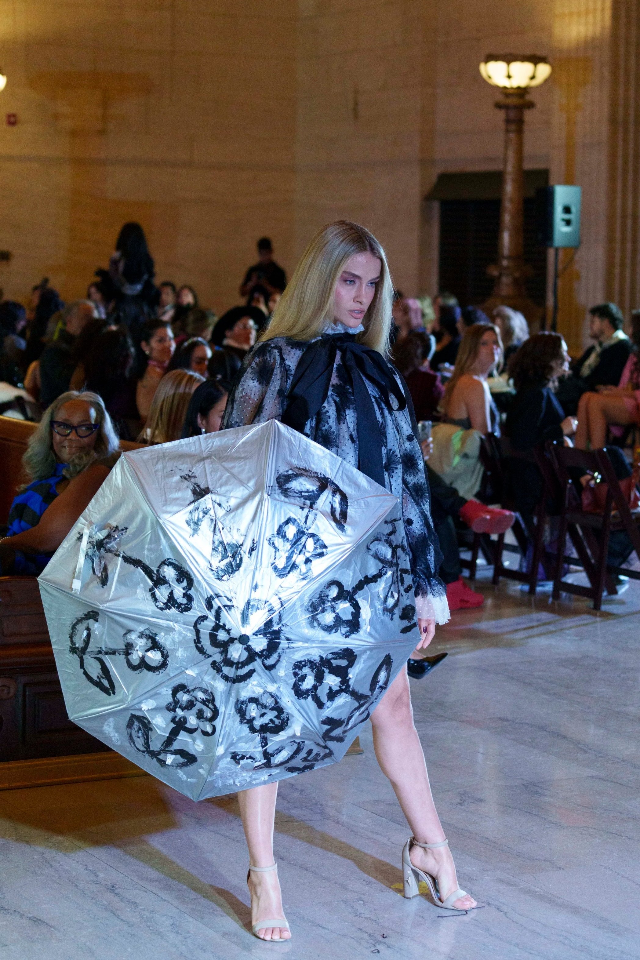 A fashion model is walking on a runway, holding a large umbrella decorated with black floral patterns. She is wearing high heel sandals and a sheer dress with black floral designs. The audience is seated on both sides of the runway, with some onlooke