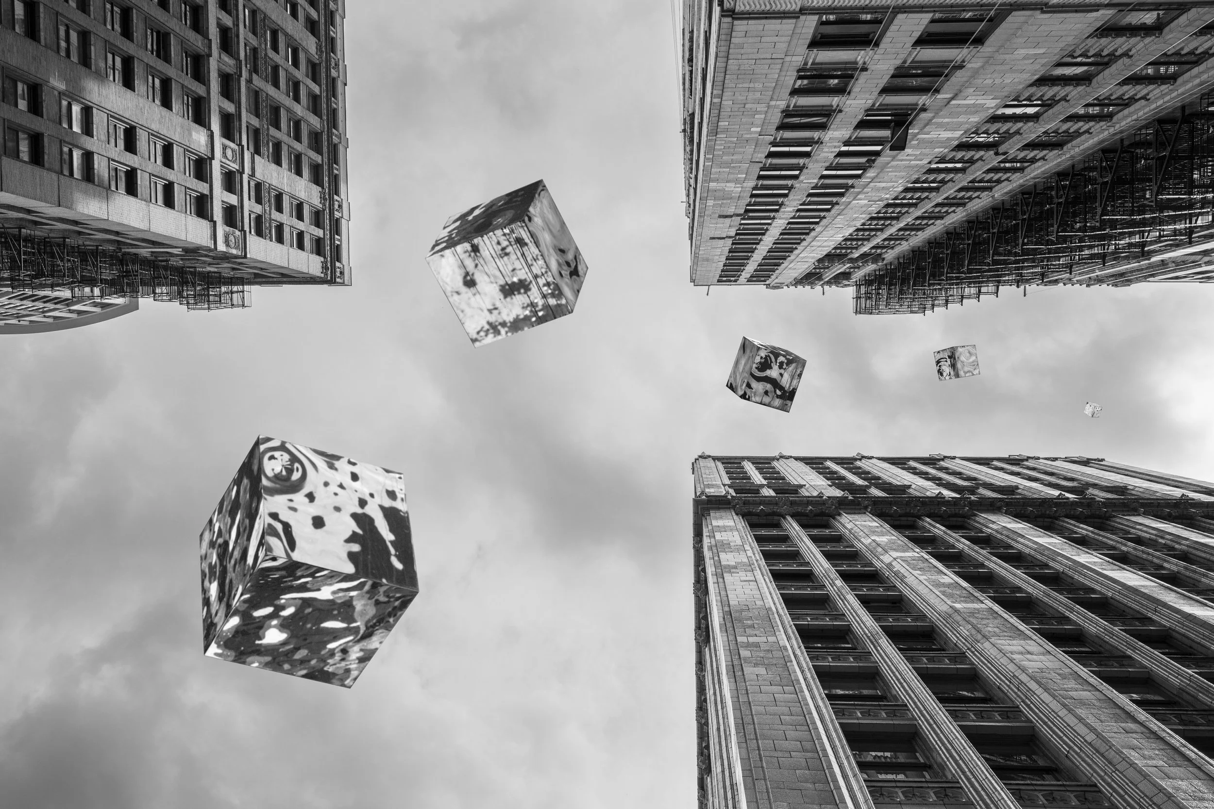 Black and white photo of tall buildings viewed from below with decorative floating cubes in the sky.