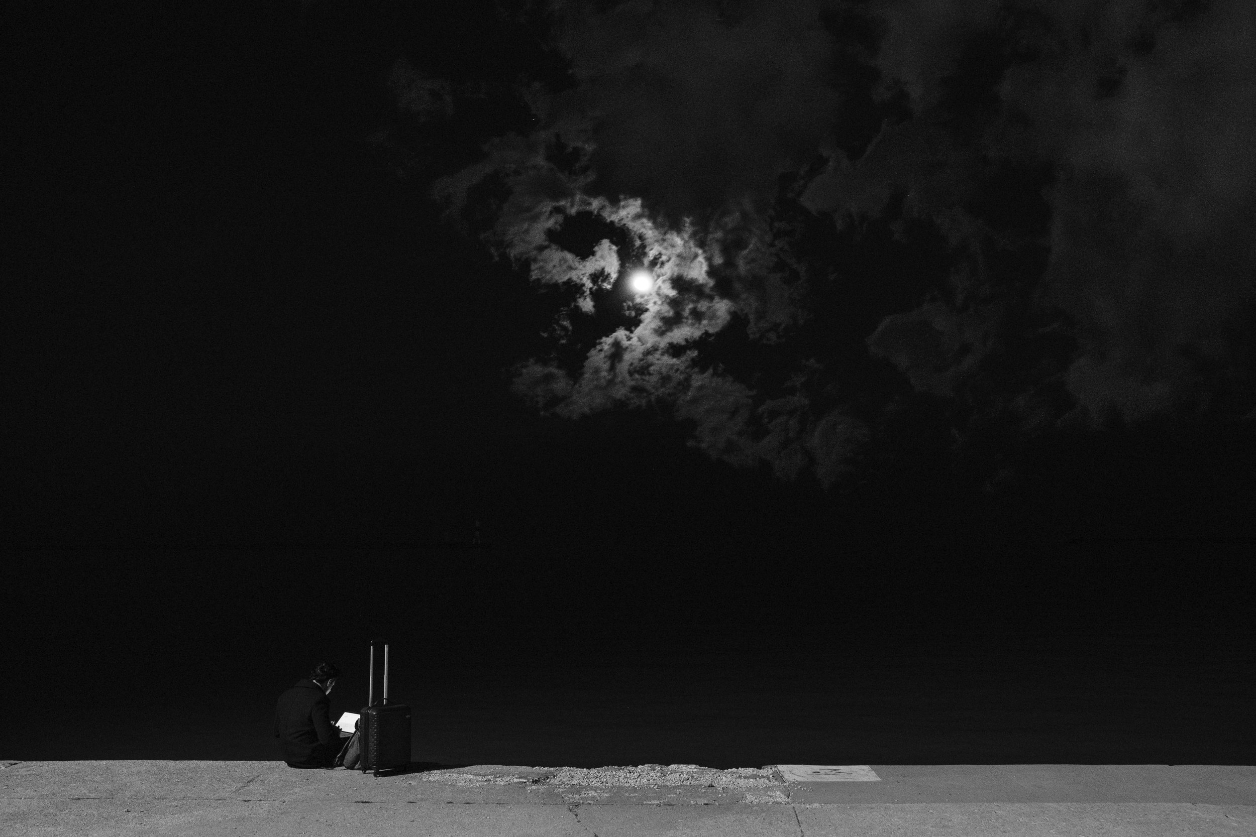 A person sitting on the ground at night, facing away from the camera, with a moon shining through dark, cloudy sky in the background.