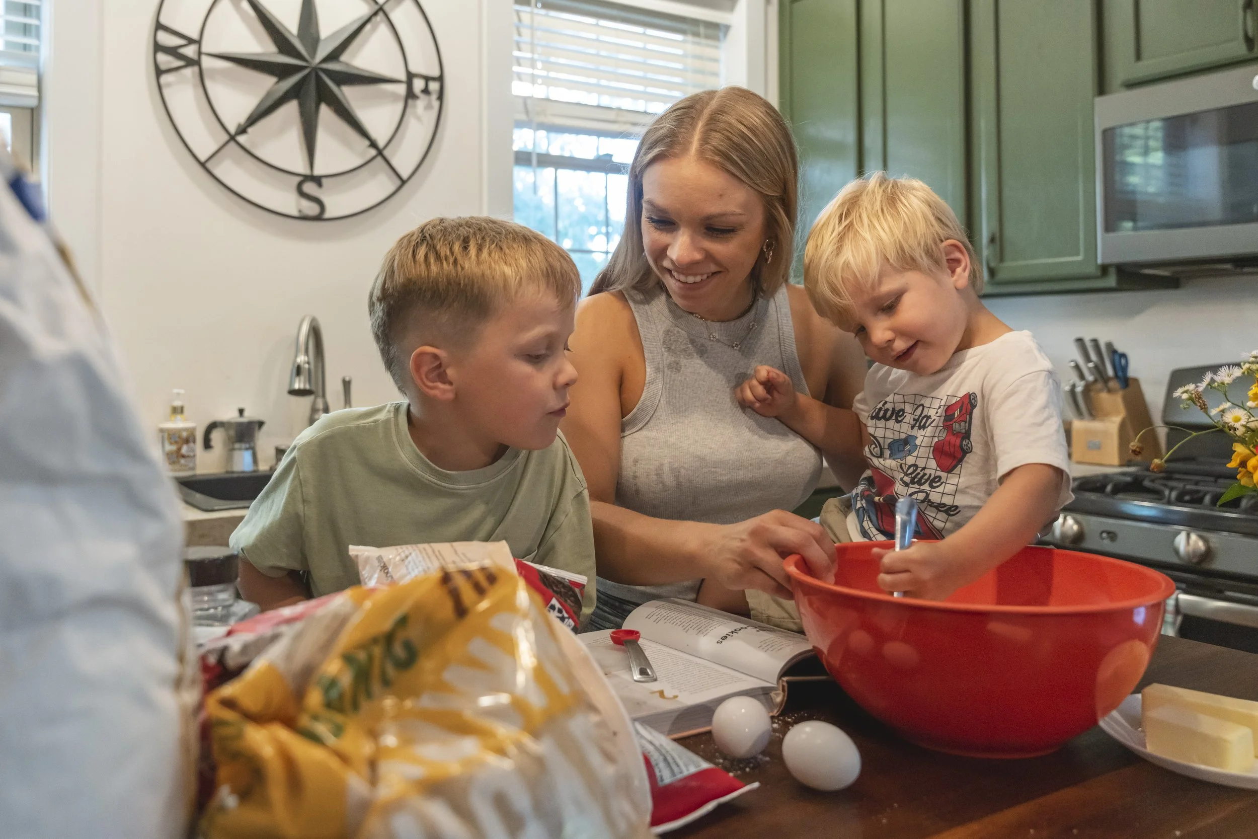 A woman with two young boys preparing food in a kitchen, with a large clock on the wall and green cabinets in the background.