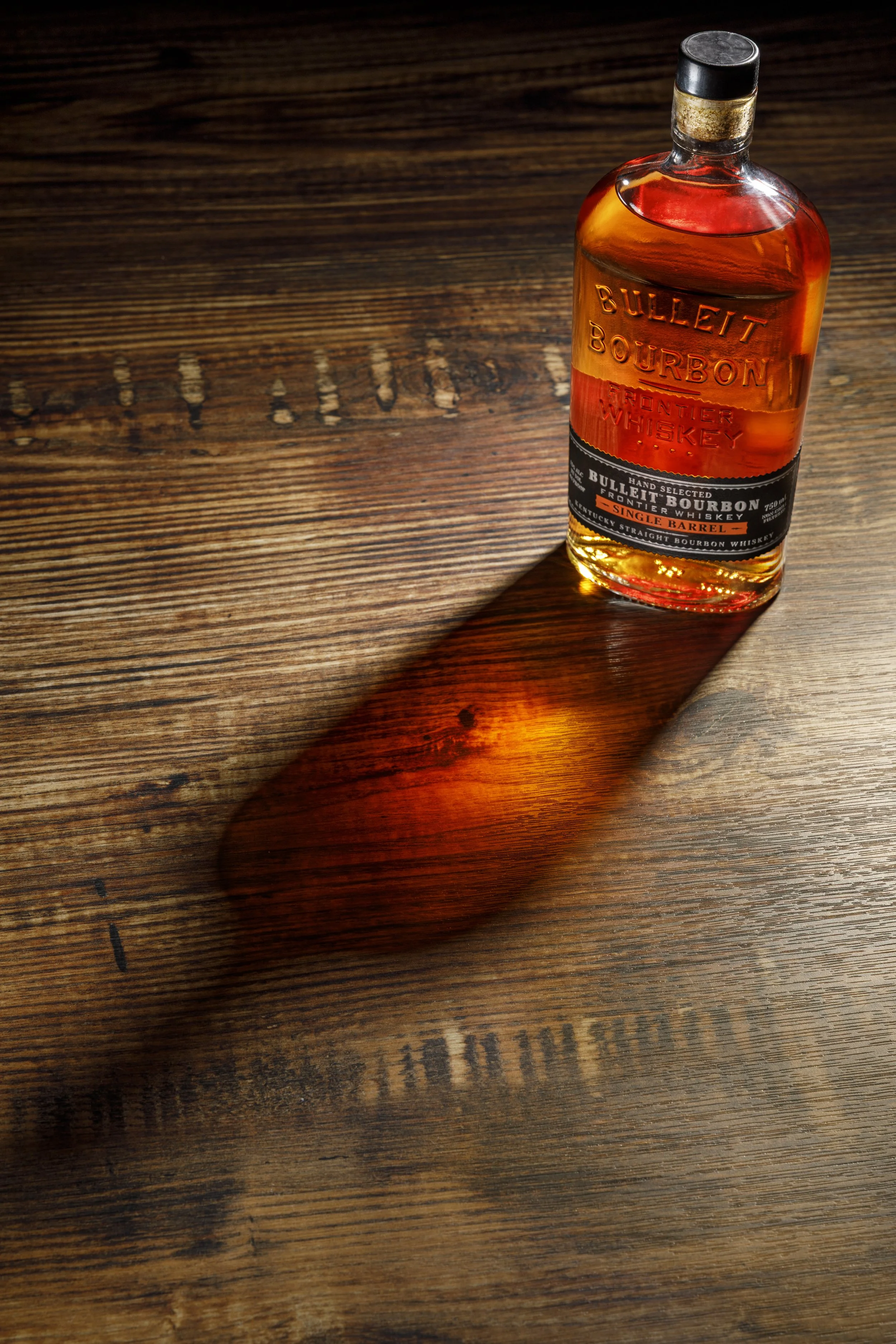 A bottle of Bulleit Bourbon whiskey on a wooden surface, with light reflecting off the bottle, casting a shadow.