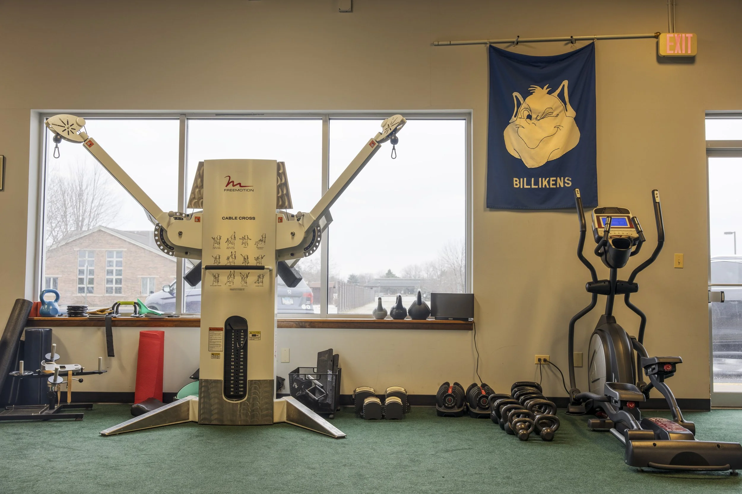 A gym with various workout equipment. In the center, there is a freemotion cable cross machine. To the right, an exercise bike and a row of dumbbells. On the left, kettlebells and resistance bands. The background features large windows with a view of
