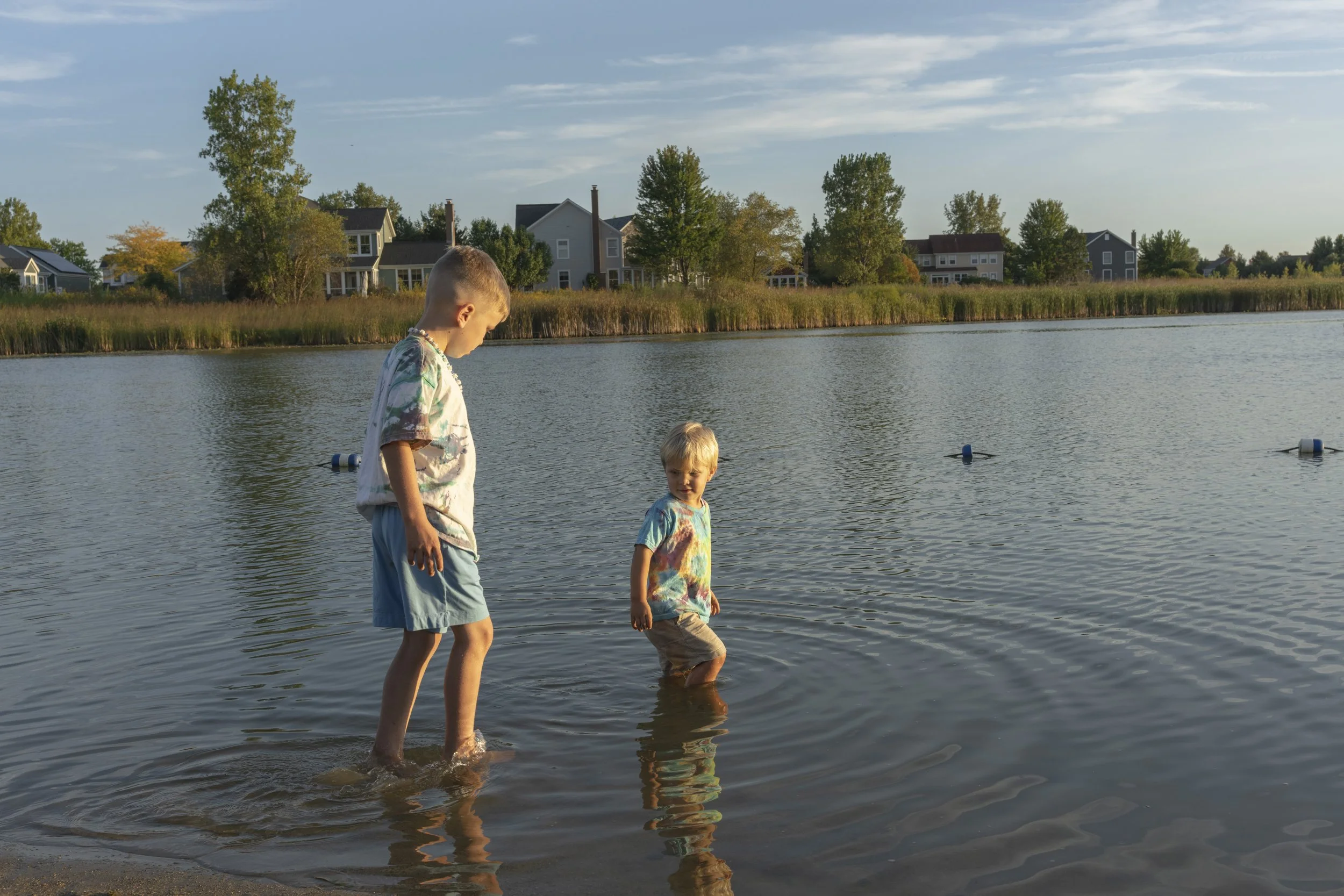 Two young boys walking in shallow water by a lake with houses and trees in the background during sunset
