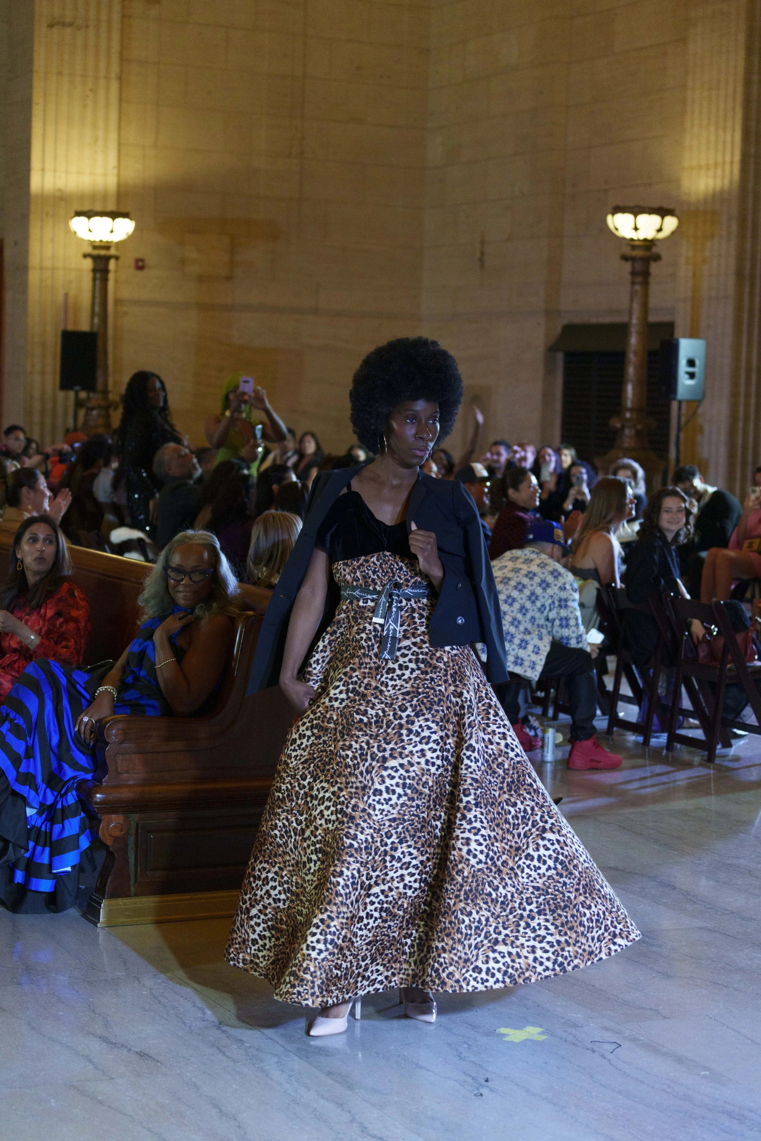 A woman with an afro hairstyle is standing in a large hall filled with seated audience members. She is wearing a leopard print skirt, white high heels, and a dark jacket draped over her shoulder. Others in the audience are taking photos or sitting an