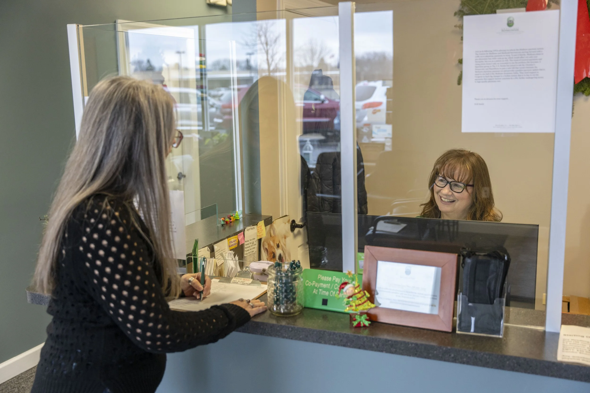 A woman with long hair and glasses at a reception counter talking to a smiling female employee behind a glass partition. The counter has paperwork, a small sign, and holiday decorations.