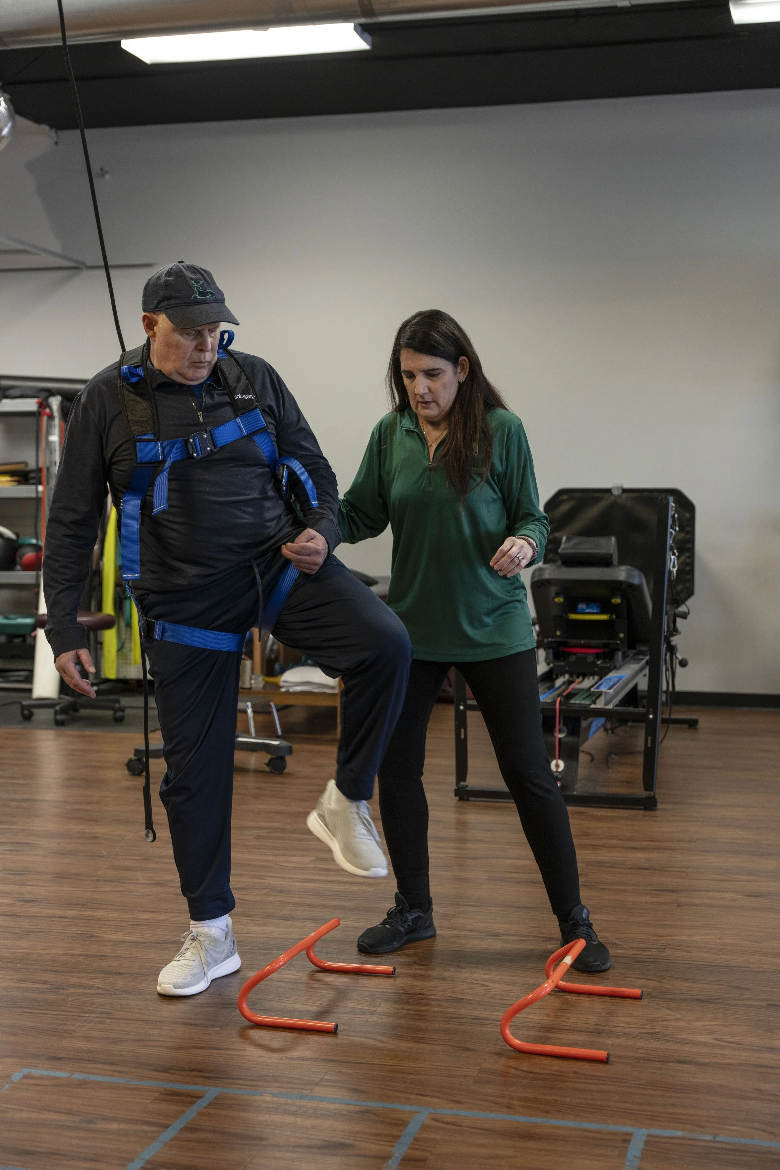 A man and a woman are in a gym, practicing agility exercises. The man is wearing a black jacket, black cap, and white sneakers, and is holding onto a resistance band with one leg raised. The woman, in a green shirt and black pants, is helping by guid