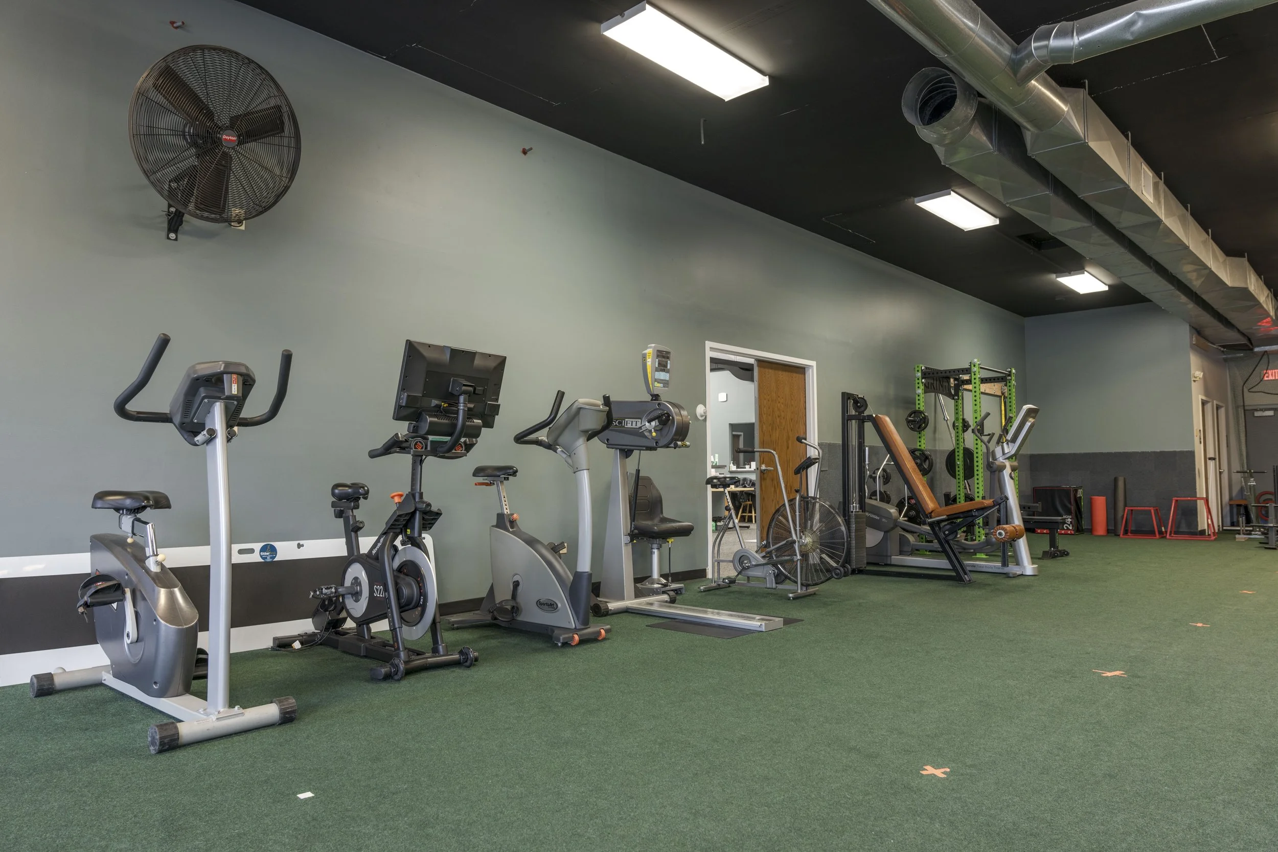 An indoor gym with exercise equipment including stationary bikes, a rowing machine, and weightlifting rack on green turf flooring, with a high ceiling and industrial ductwork.