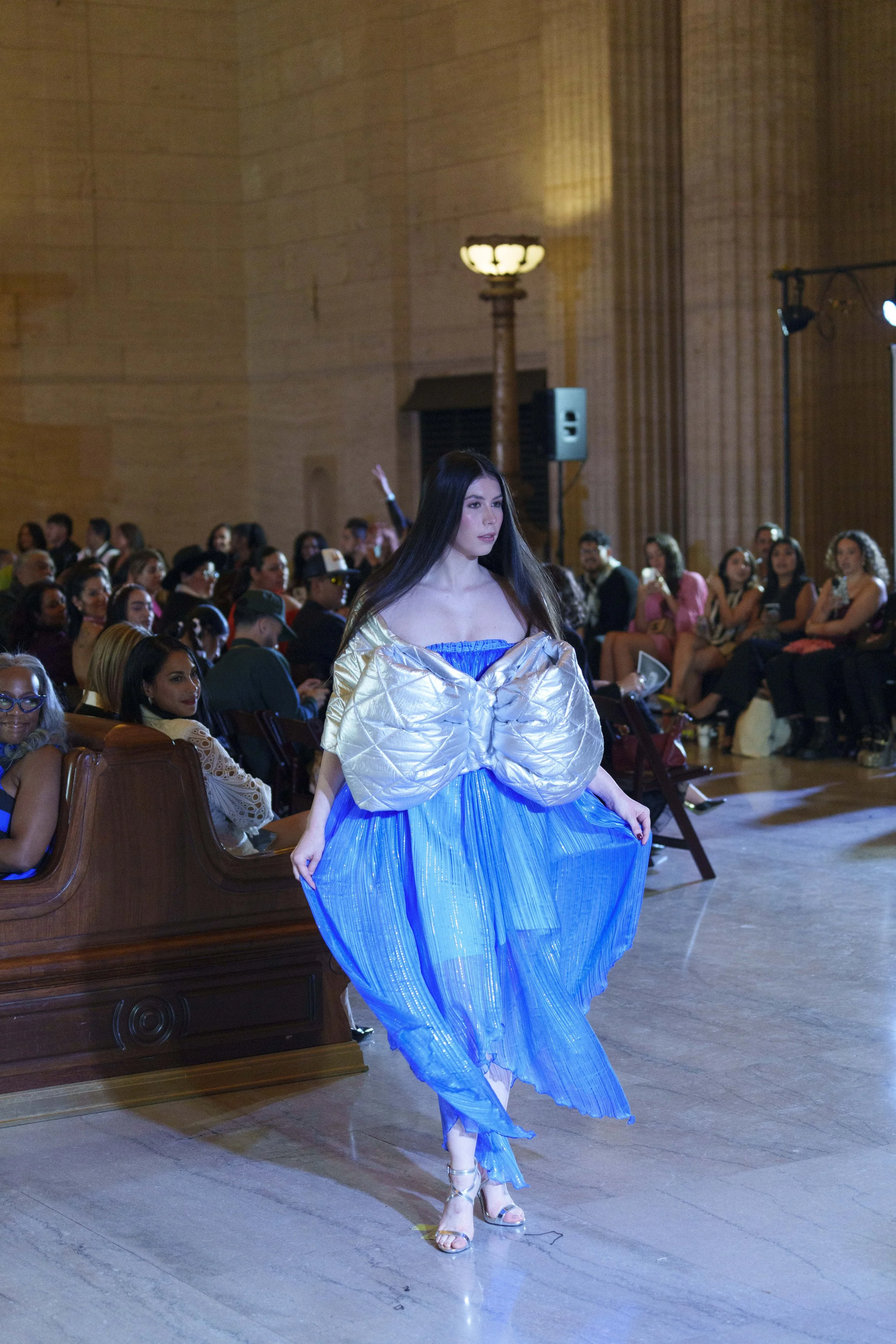 A young woman walking on a runway during a fashion show, wearing a sheer blue dress with a puffy golden top, in a room filled with seated audience members watching her.