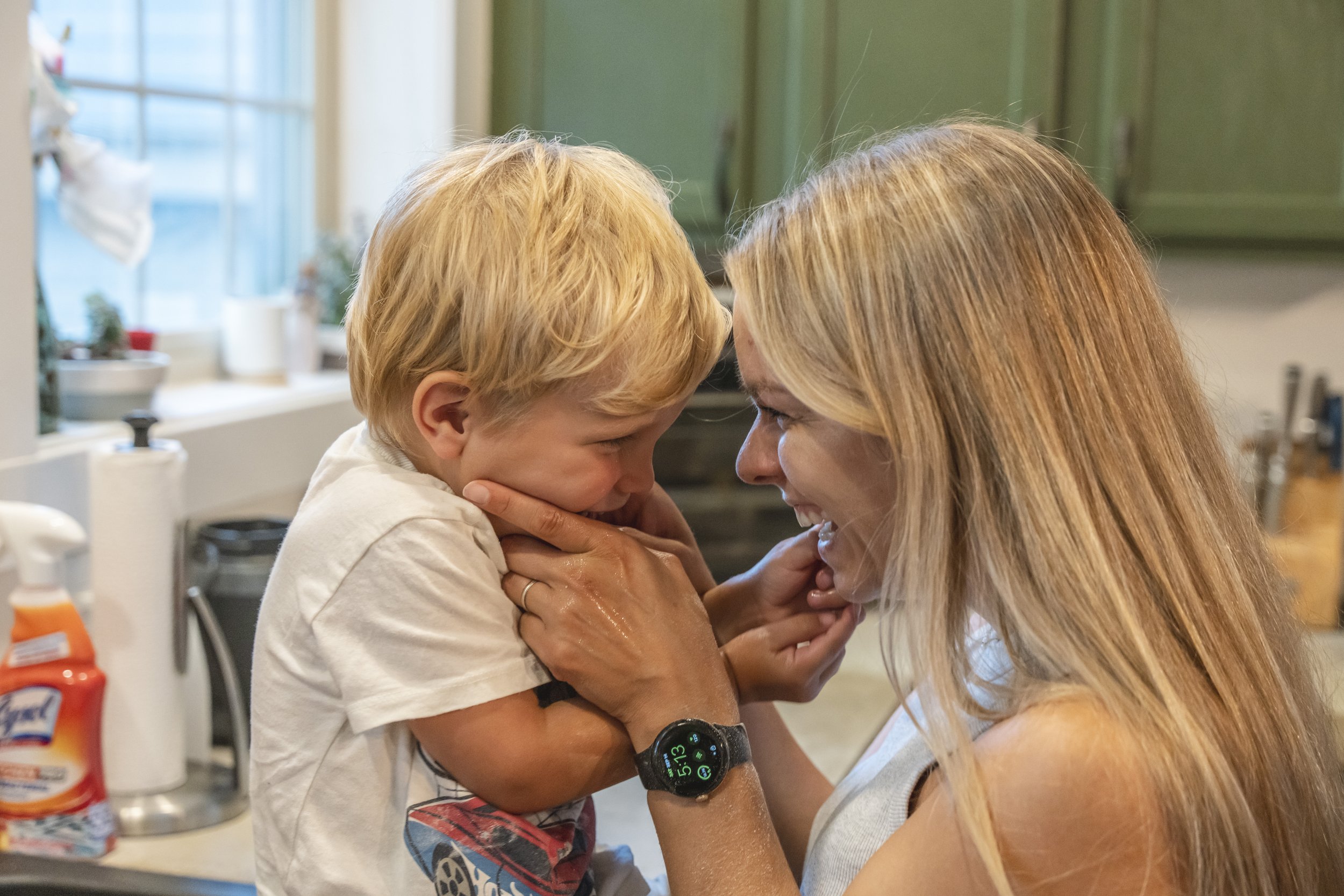 A woman and a young boy with blonde hair are face-to-face, touching foreheads and smiling in a kitchen.