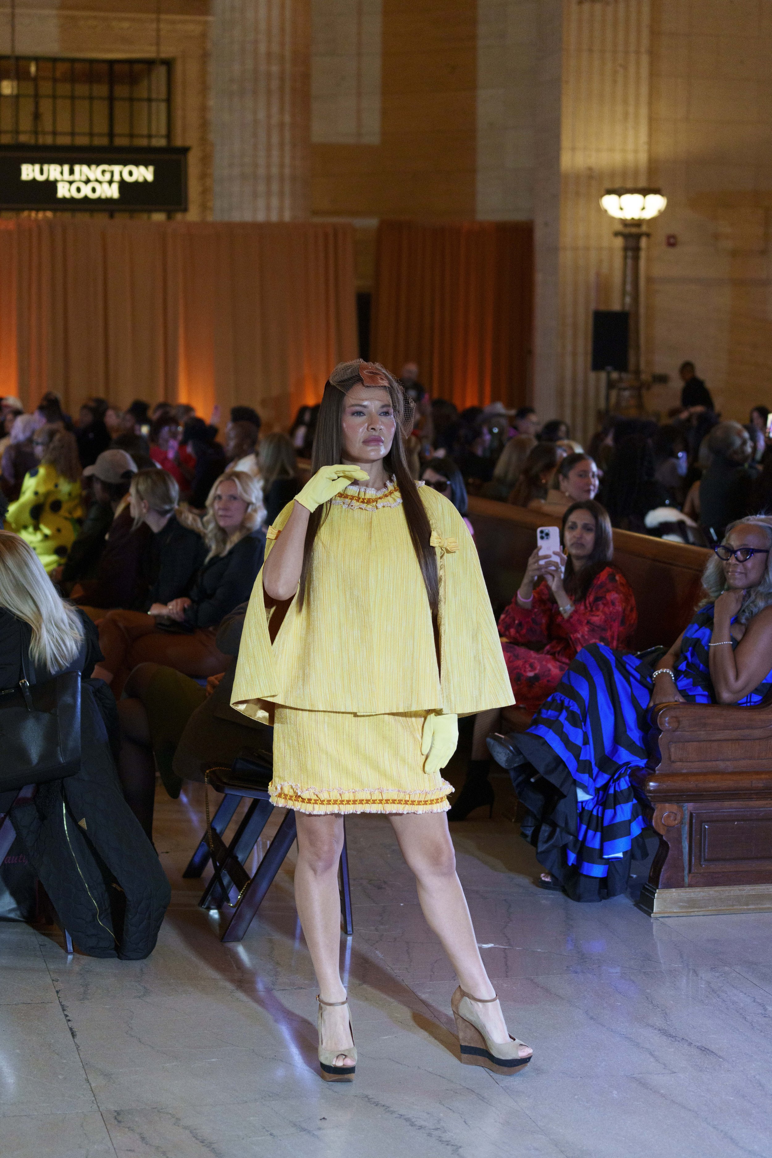 A person modeling in a yellow dress with matching gloves, standing on a runway at a fashion show, with audience members seated on wooden pews and holding smartphones in the background.