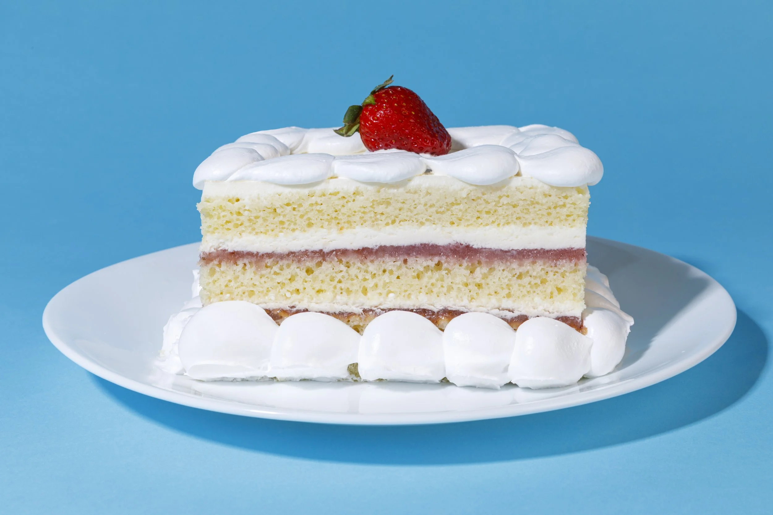 A slice of strawberry shortcake on a white plate, topped with a whole strawberry and white whipped cream, against a blue background.