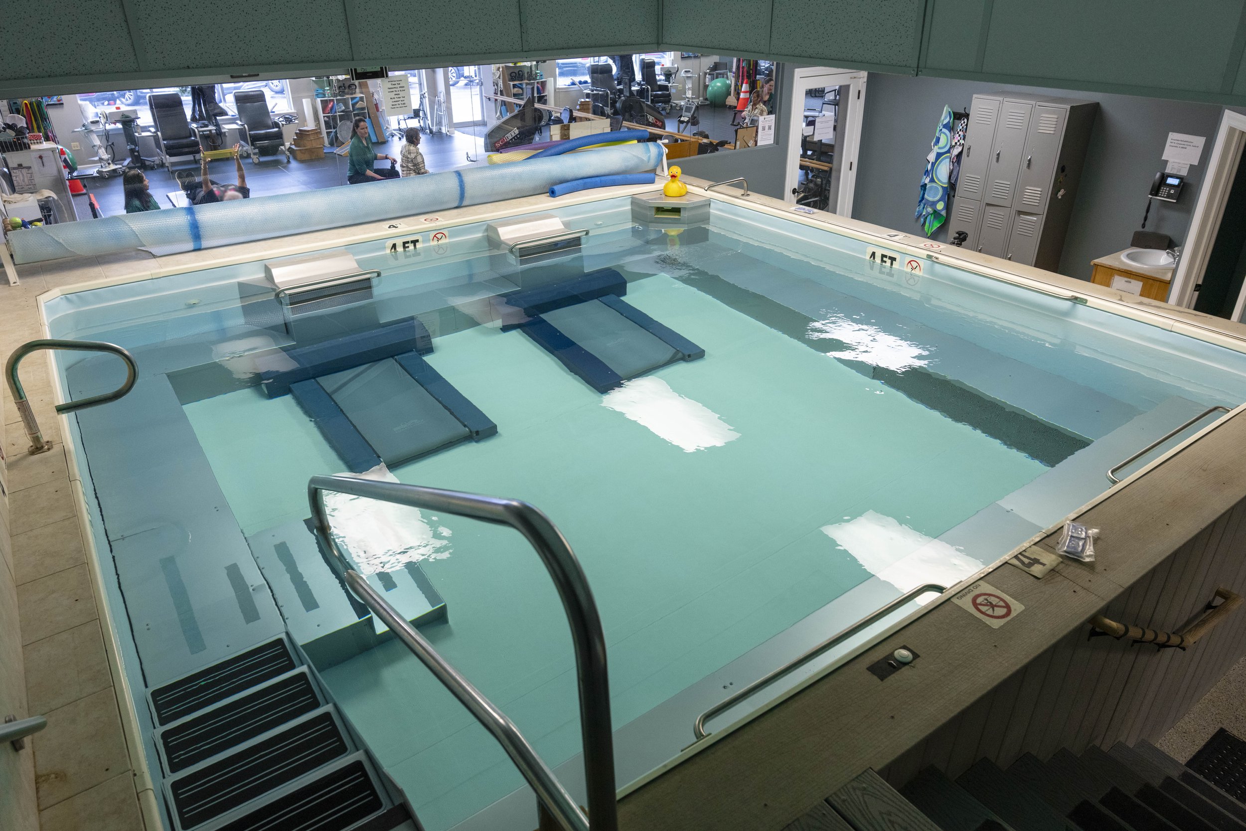 Indoor therapy pool with a rubber duck, blue foam floaties, and lounger seats submerged. Observation room with fitness equipment and individuals, including a person practicing exercises, visible through a large window.