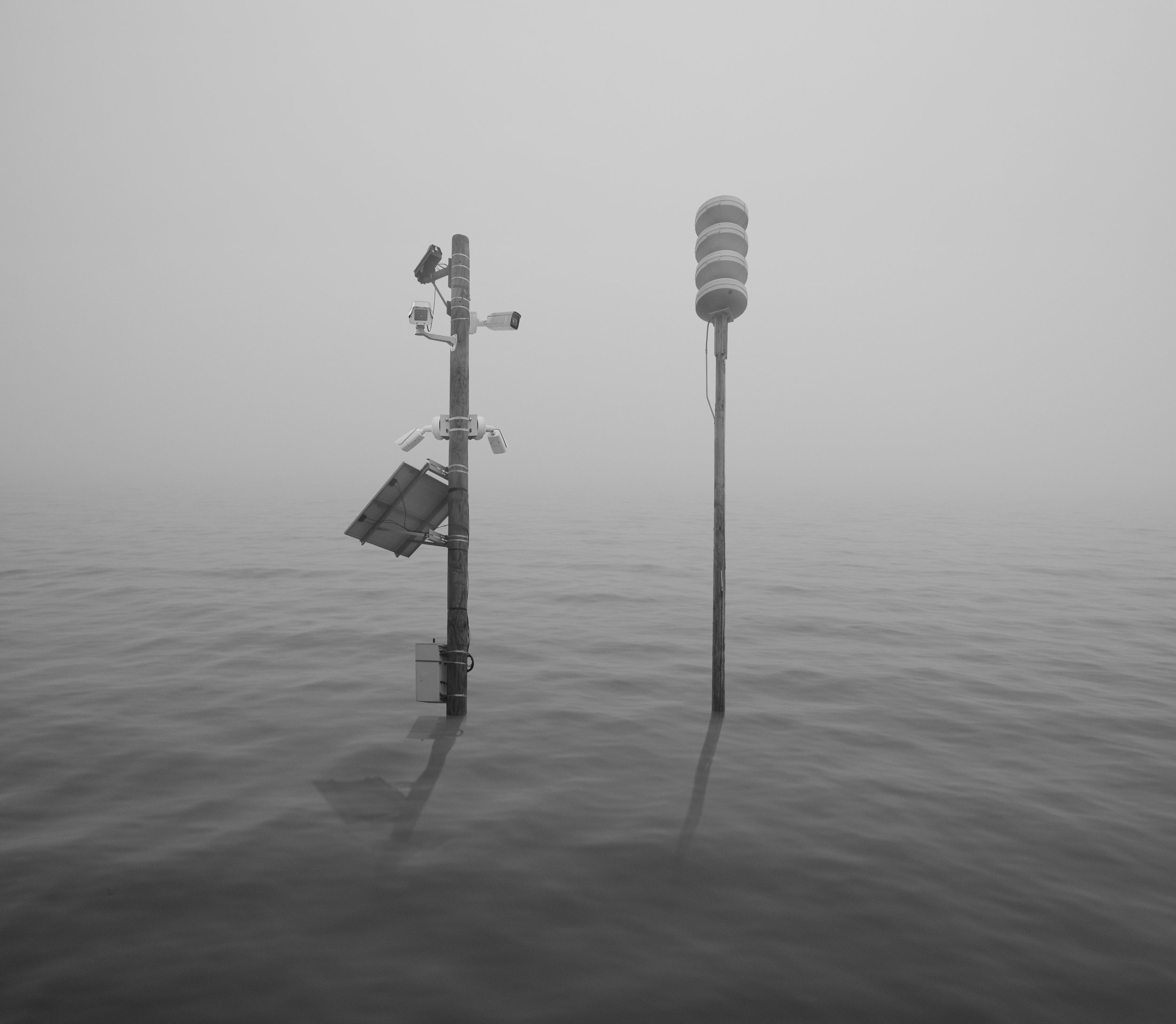 Two navigation beacons stand in the ocean, casting reflections on the water, with a hazy, overcast sky overhead.