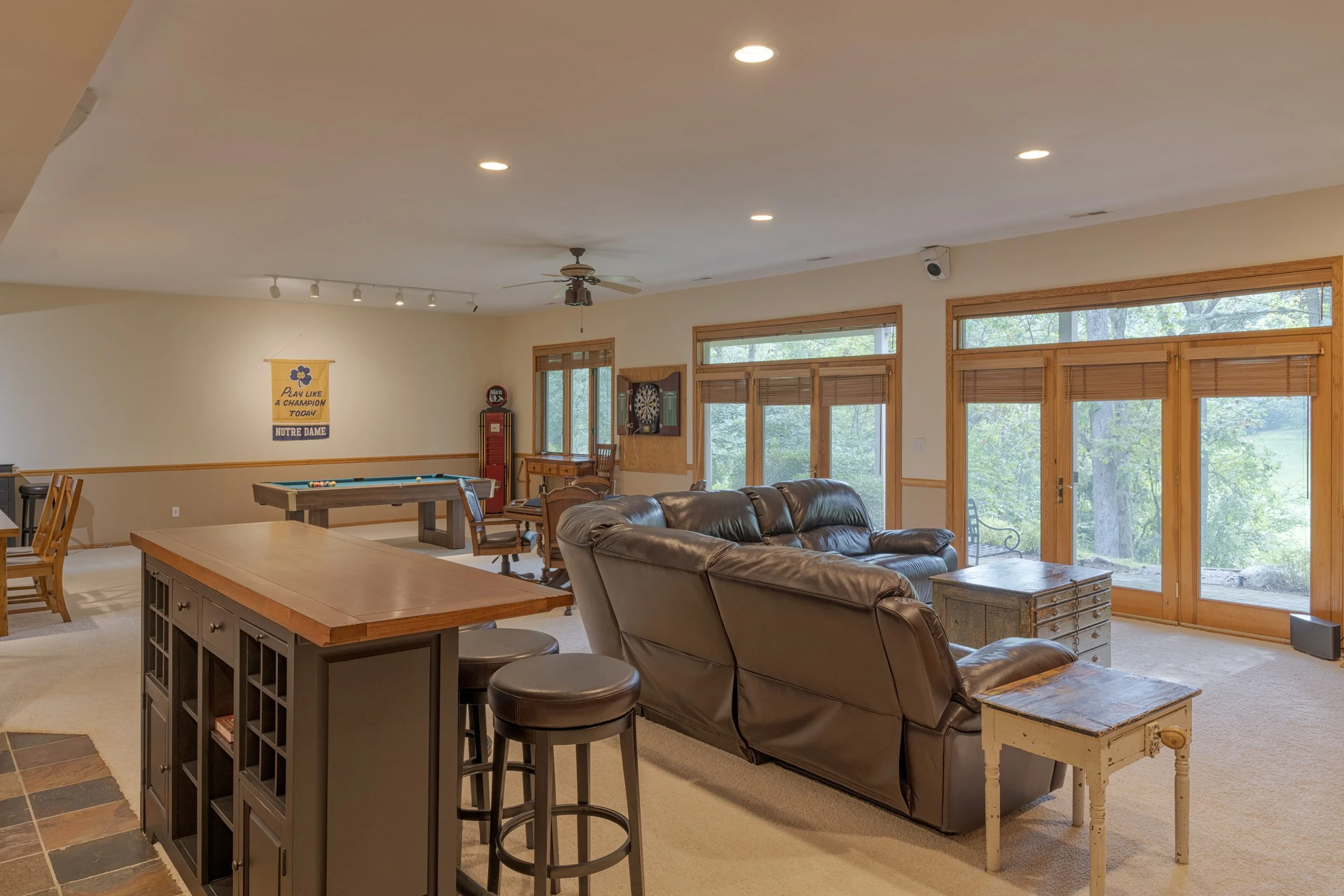 Living room with large windows and wood frames, a brown leather sofa, a wooden coffee table, a small white side table, a dartboard, a ceiling fan, and a banner on the wall that reads 'Play Like a Champion Today Notre Dame'.