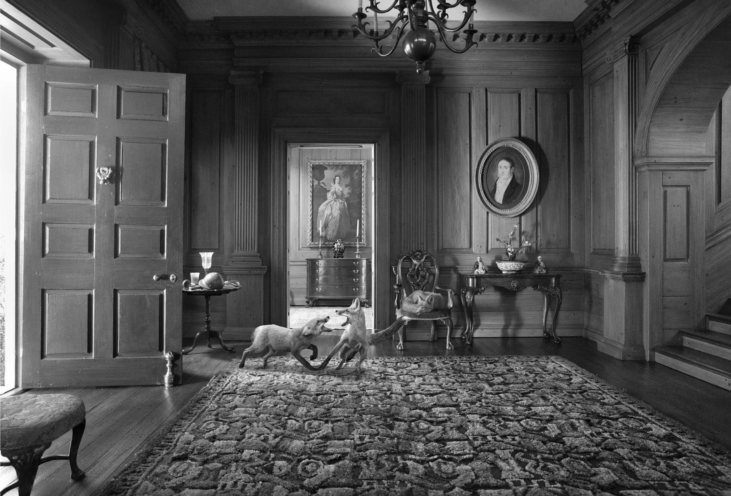 A hallway with wooden paneled walls, ornate crown molding, and a chandelier hanging from the ceiling. There are framed portraits on the walls, a side table with a bowl and statues, and four dogs playfully interacting on a patterned rug. An open door 