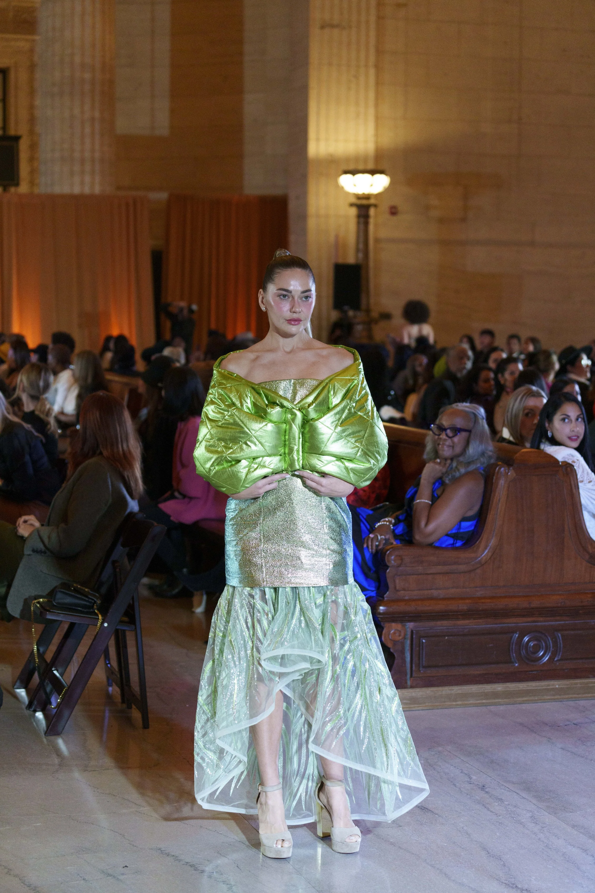 A female model walking on a runway at a fashion show, wearing a shiny green and gold gown with puffed sleeves and a sheer, patterned skirt, in a large wooden hall filled with seated audience members.