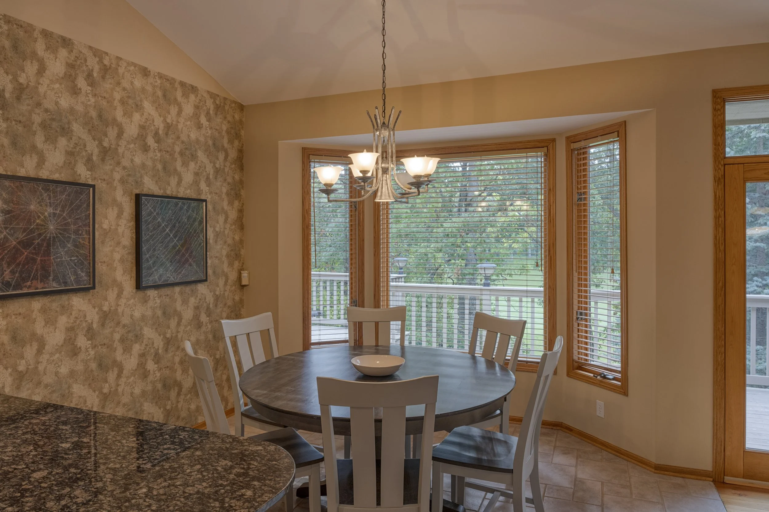 A dining area with a round table surrounded by white chairs, a bowl in the center, large windows with wooden blinds, a chandelier hanging from the ceiling, and artwork on a textured wall.
