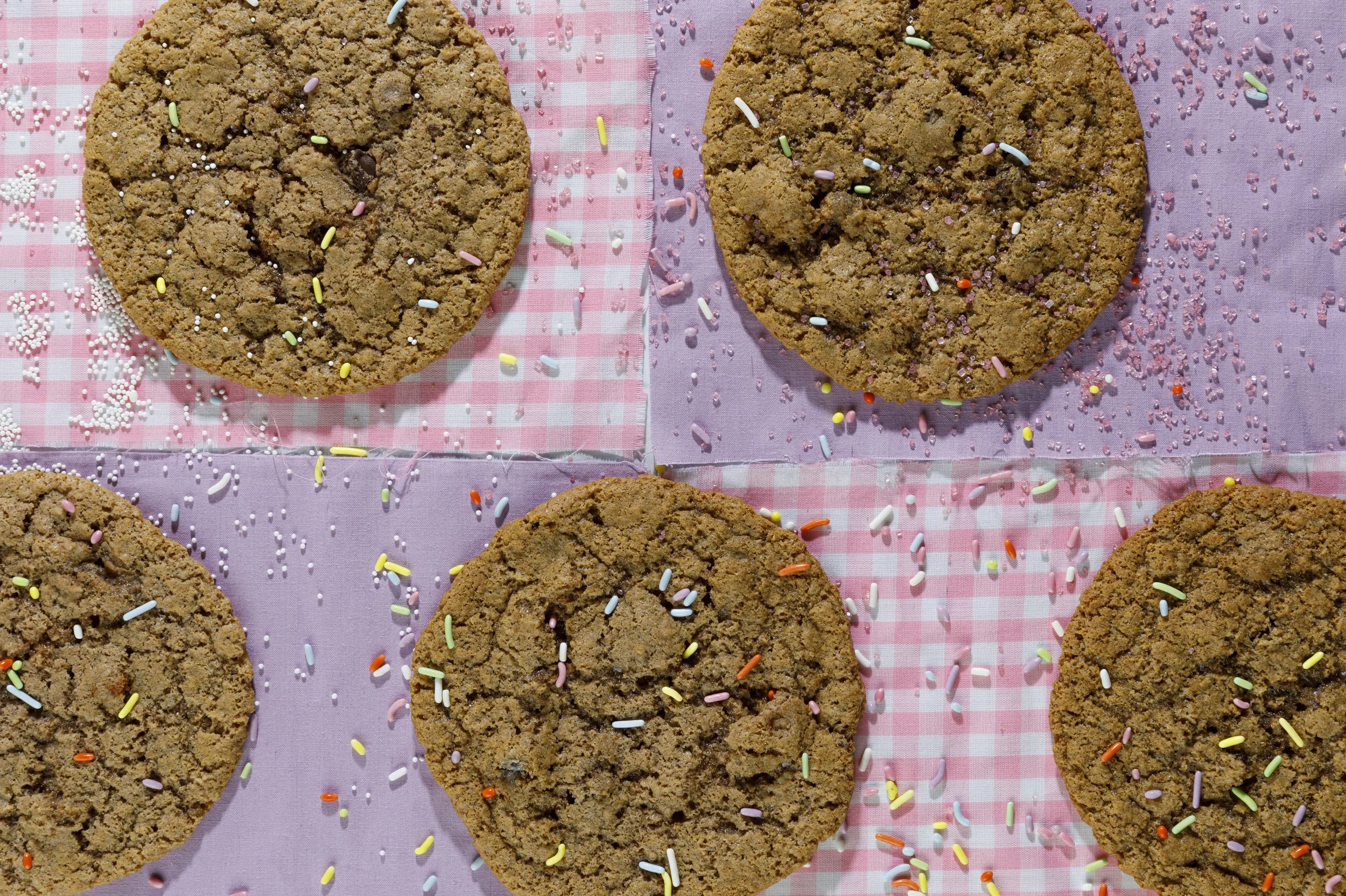 Close-up view of several large chocolate chip cookies with colorful sprinkles on a pink and white checkered cloth.