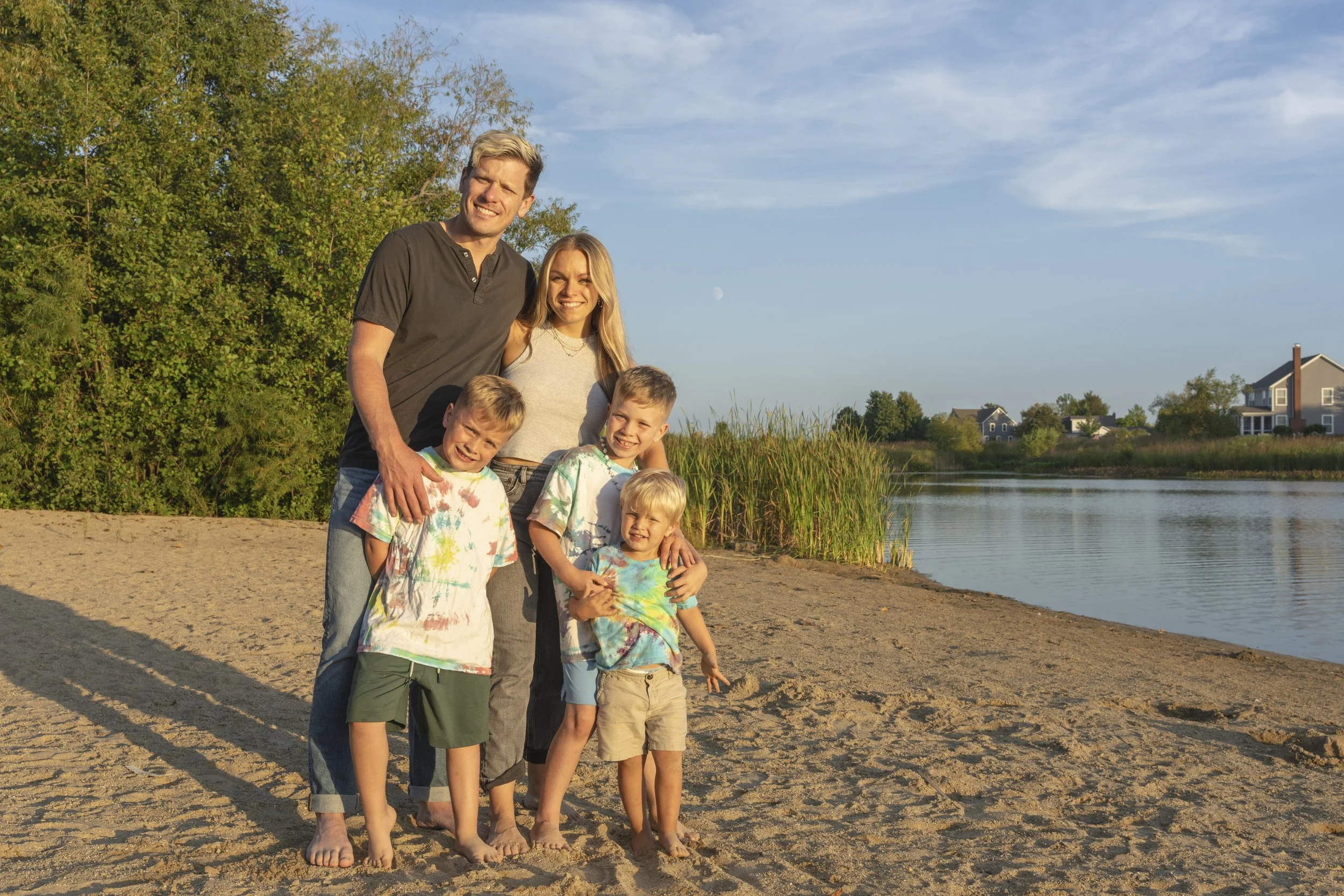 A family with four children and two adults standing together on a sandy beach by a lake, smiling and posing for a photo during sunset, with houses and trees in the background.