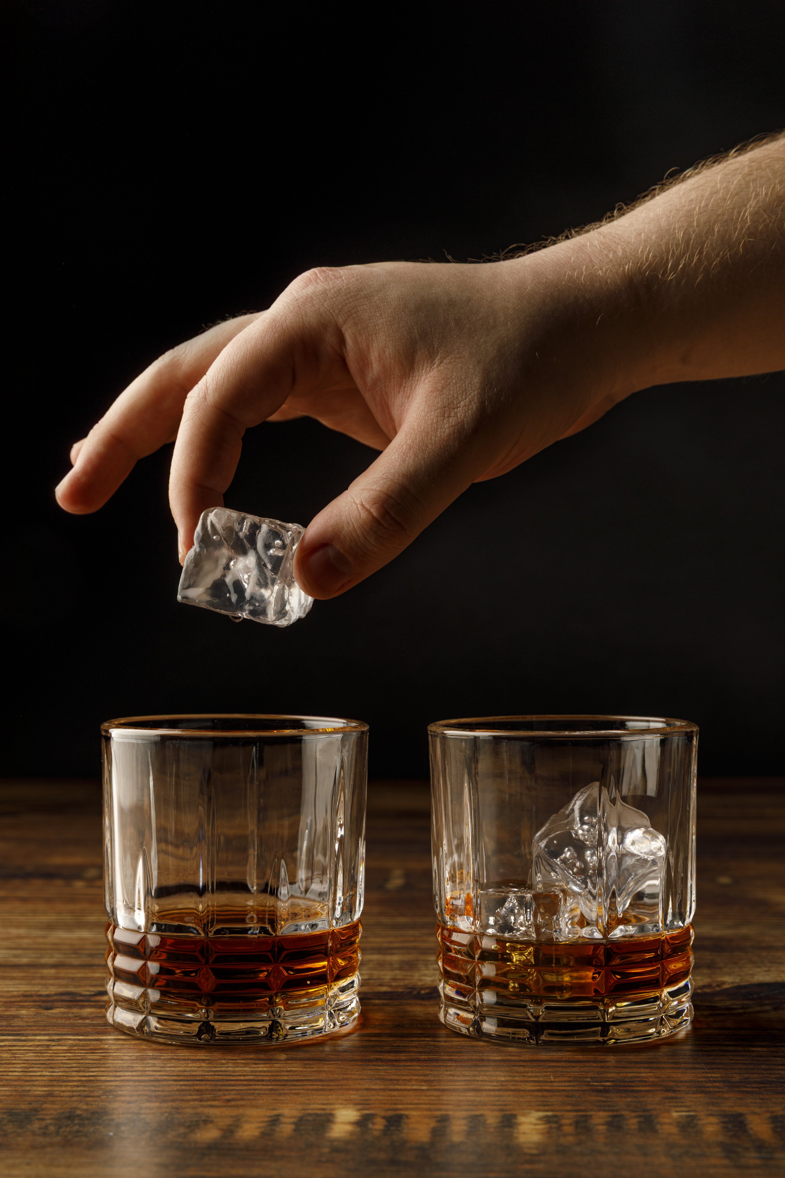 A person's hand placing a large ice cube into a glass of whiskey, with a second glass of whiskey and ice in the foreground on a wooden surface against a black background.