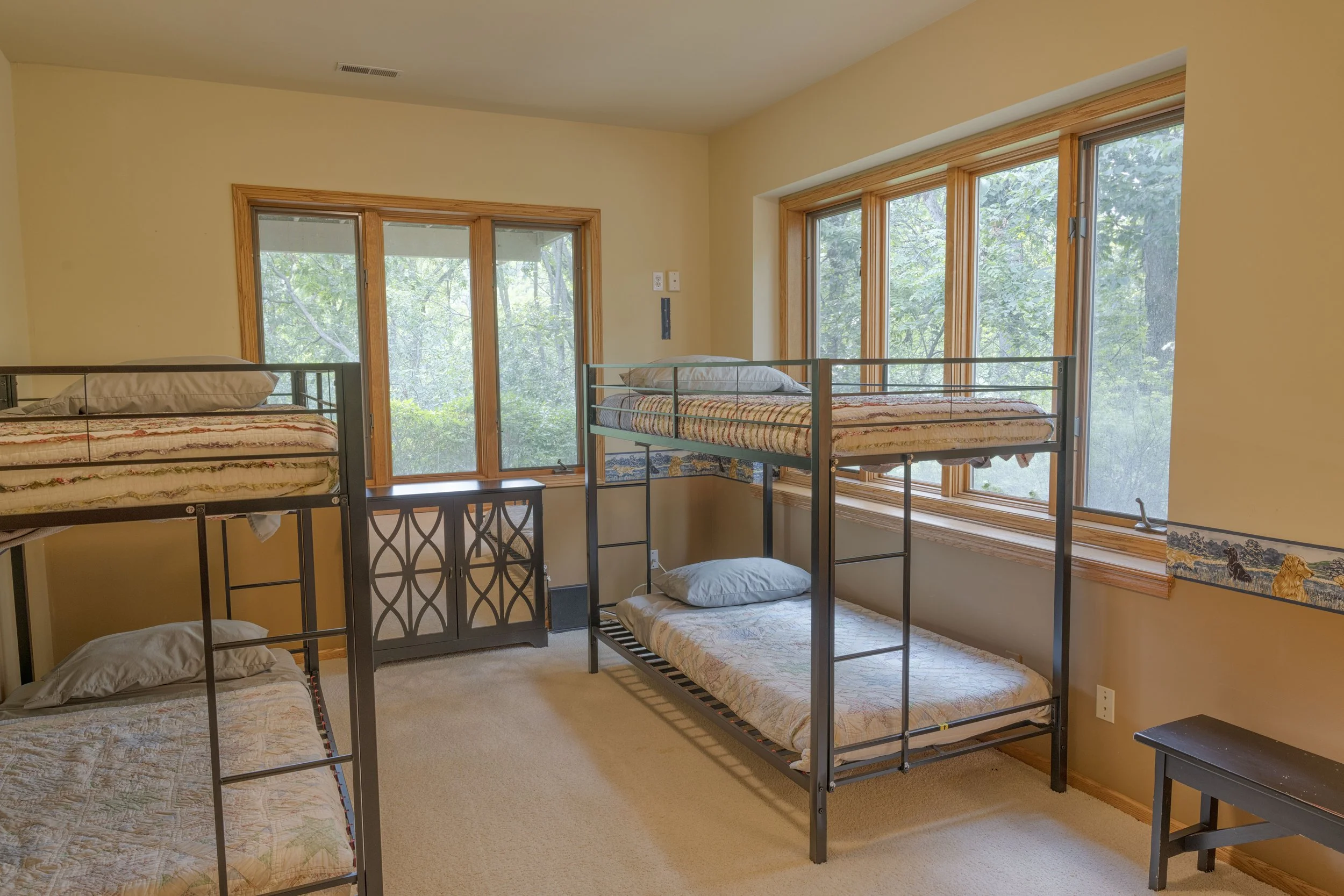 Room with multiple metal bunk beds, large windows showing green trees outside, beige walls, and small black side tables.