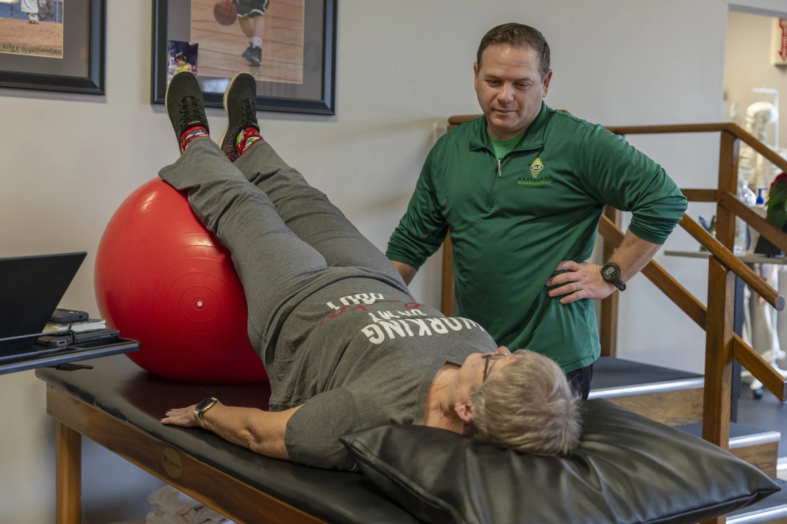 A man lying on a therapy table with his legs elevated on a large red exercise ball, engaging in physical therapy or rehabilitation, while a healthcare professional stands nearby observing.