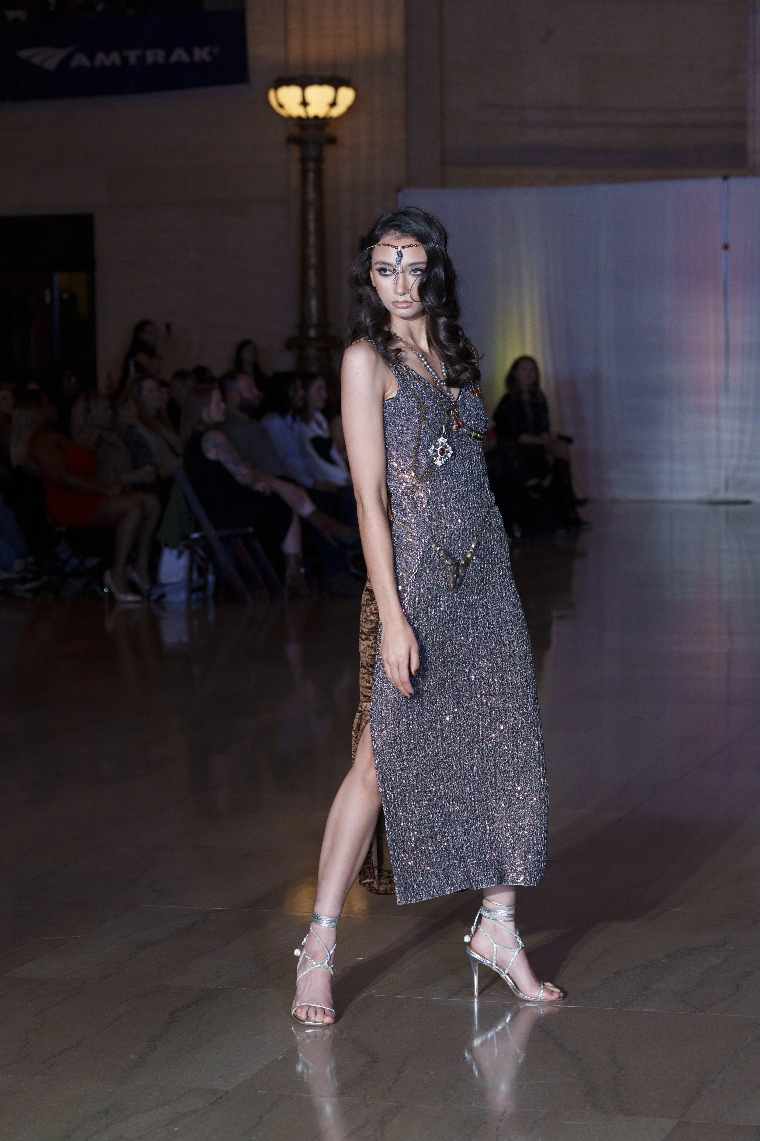 A woman walking the runway during a fashion show in a silver, shiny, slit dress with high heels.