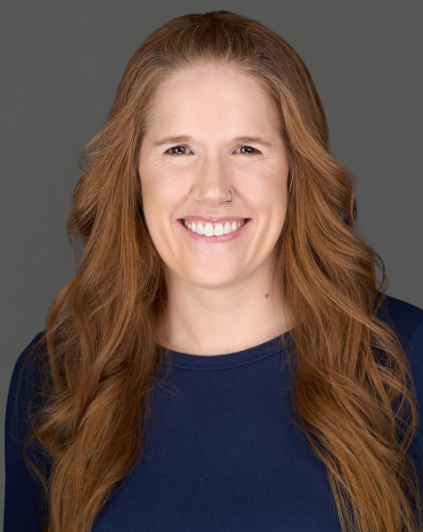 Woman with long red hair and a navy blue shirt smiling against a gray background.