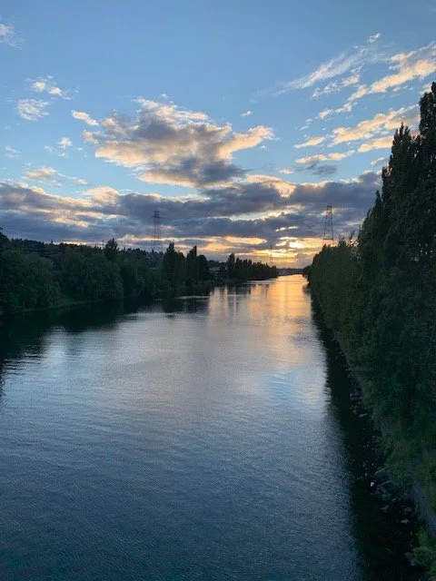 A river at sunset with partly cloudy sky, surrounded by trees and power lines in the distance.