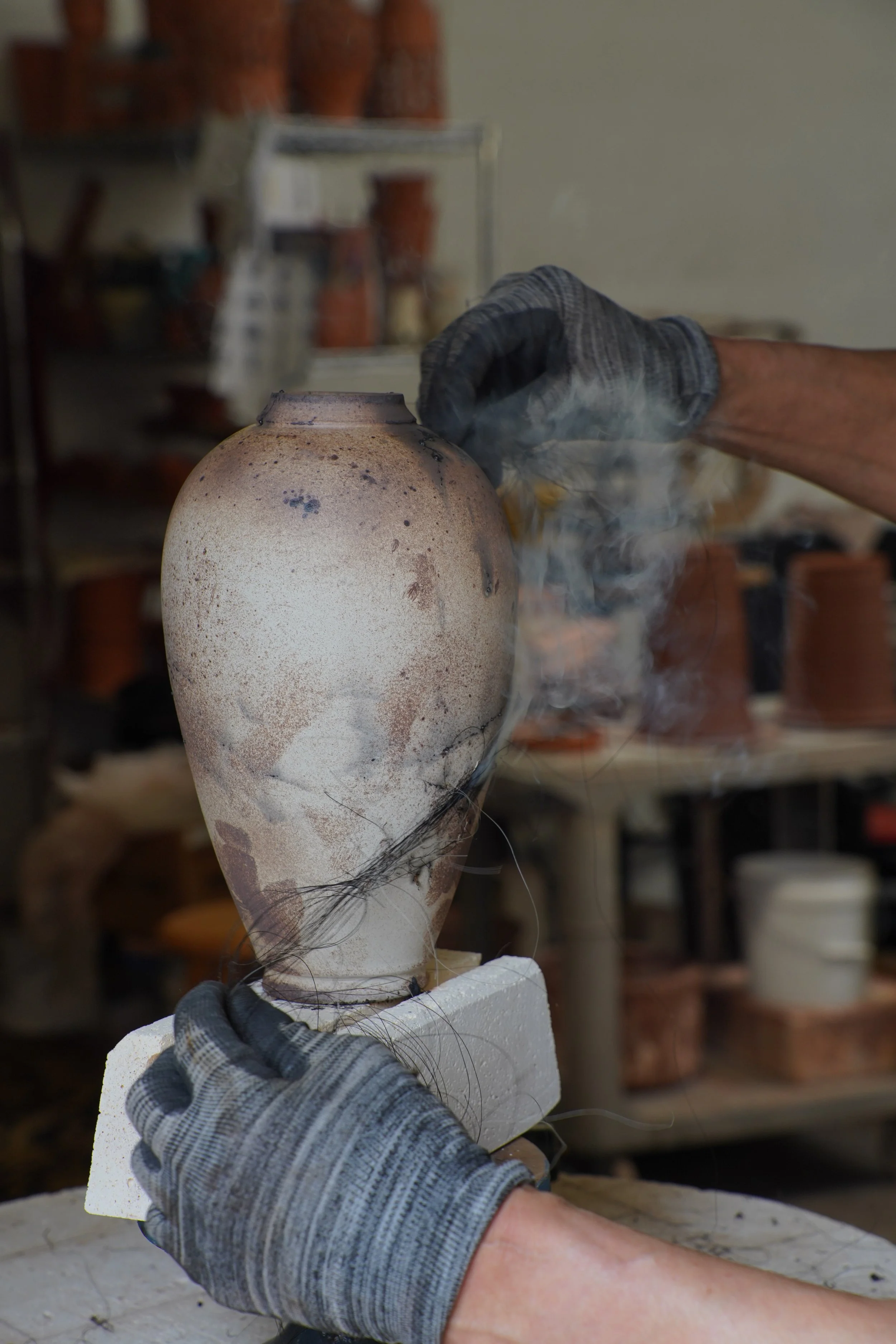 A person wearing protective gloves is placing horse hair on a burning hot ceramic vase in a pottery studio, with shelves of pottery in the background.