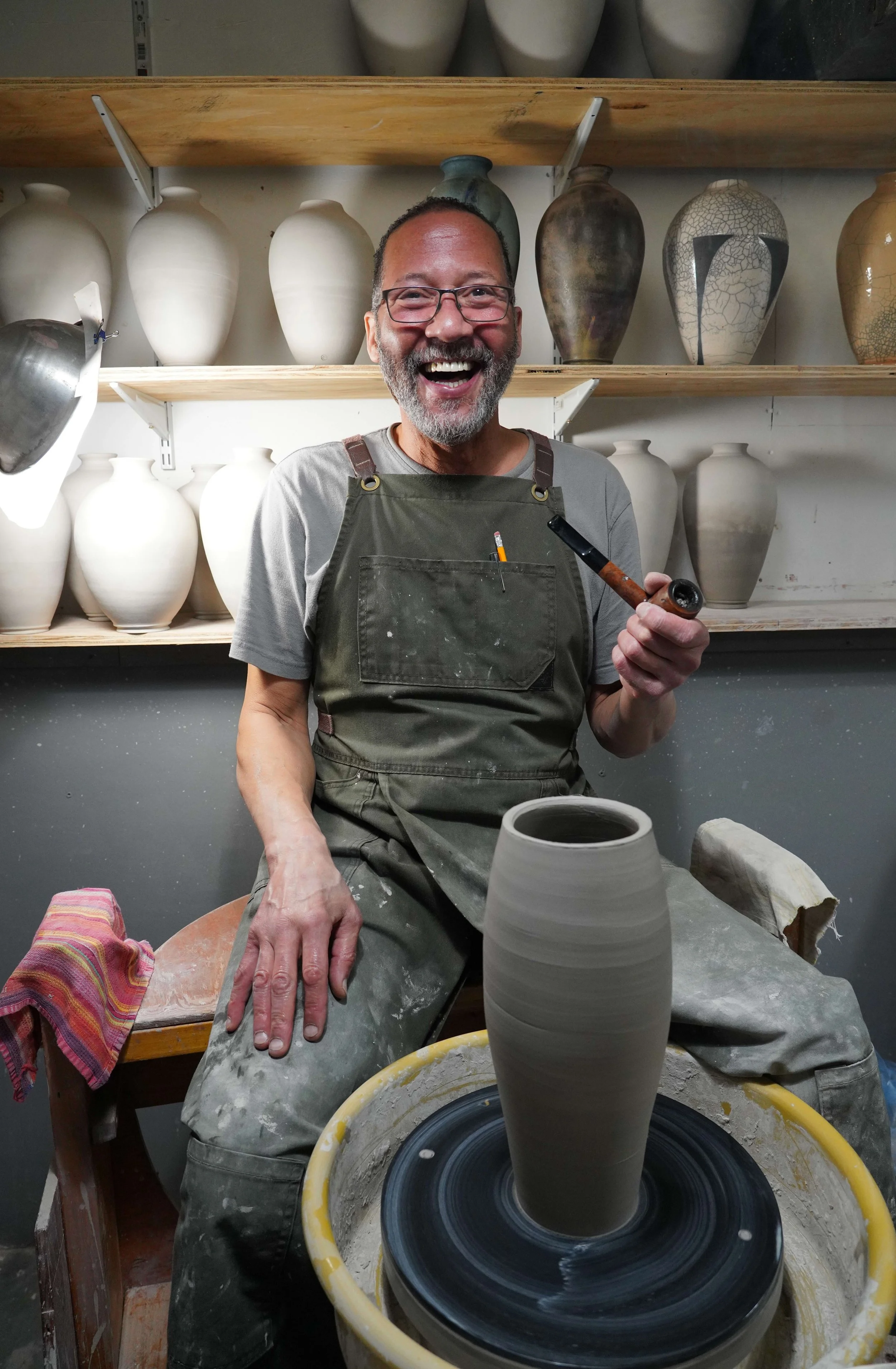 A man with glasses and a beard smiles while working on a pottery kick wheel, shaping a tall clay vase. Behind him are shelves with finished vases of various shapes and colors.