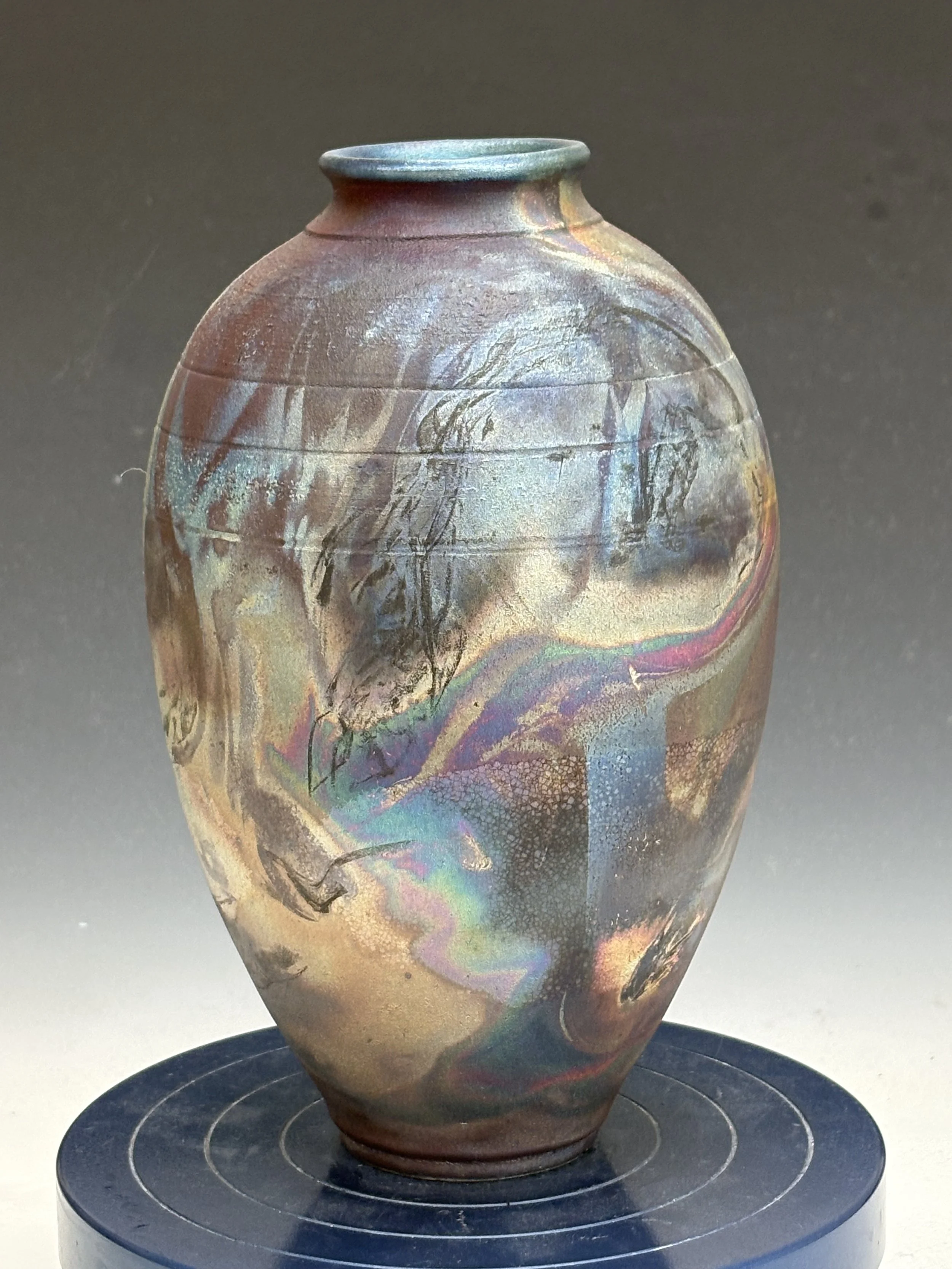 A ceramic vase with an iridescent glaze featuring swirling colors and patterns, displayed on a circular black stand.