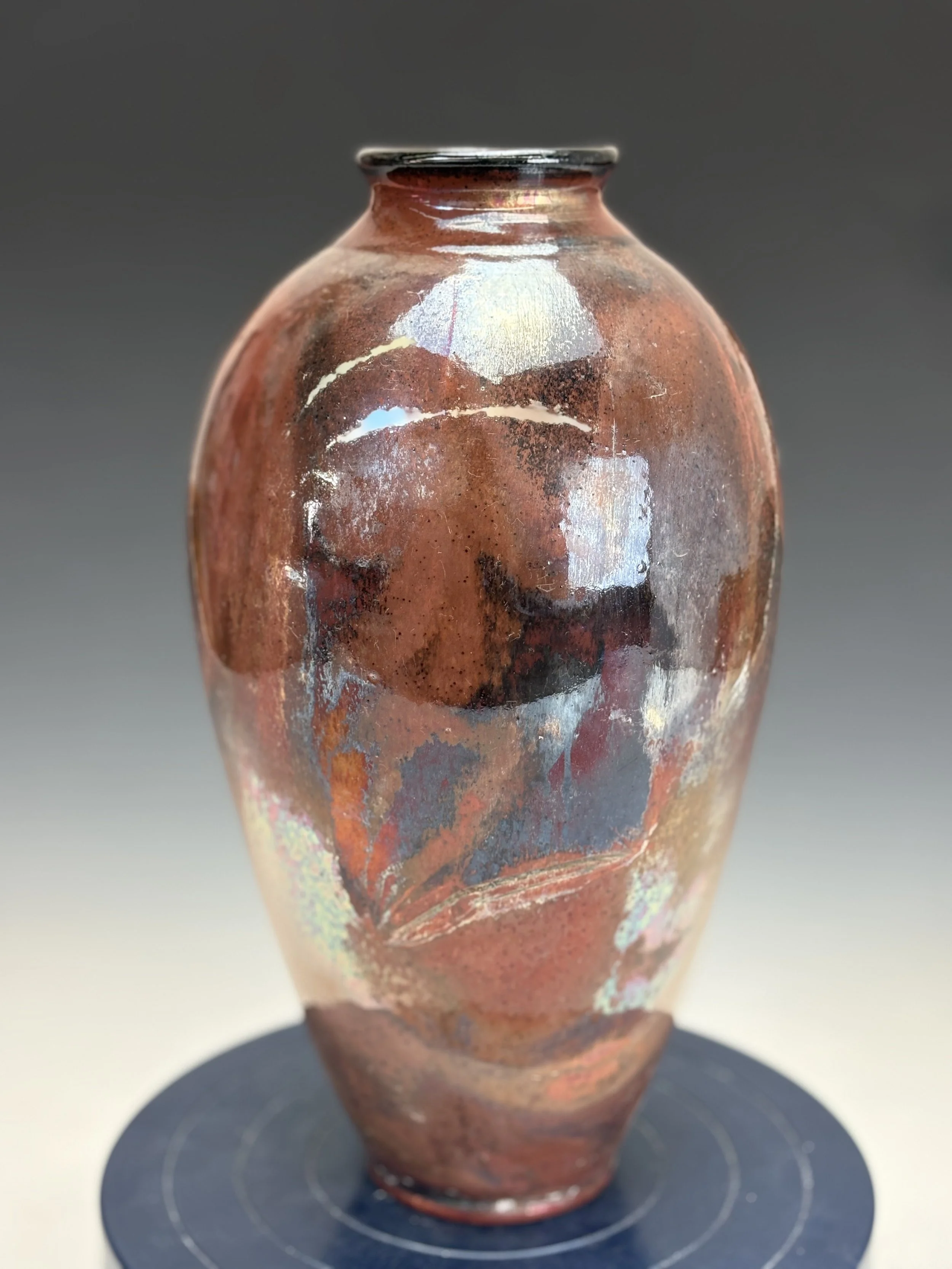 Close-up of a ceramic vase with a glossy, reddish-brown glaze, placed on a black circular stand.
