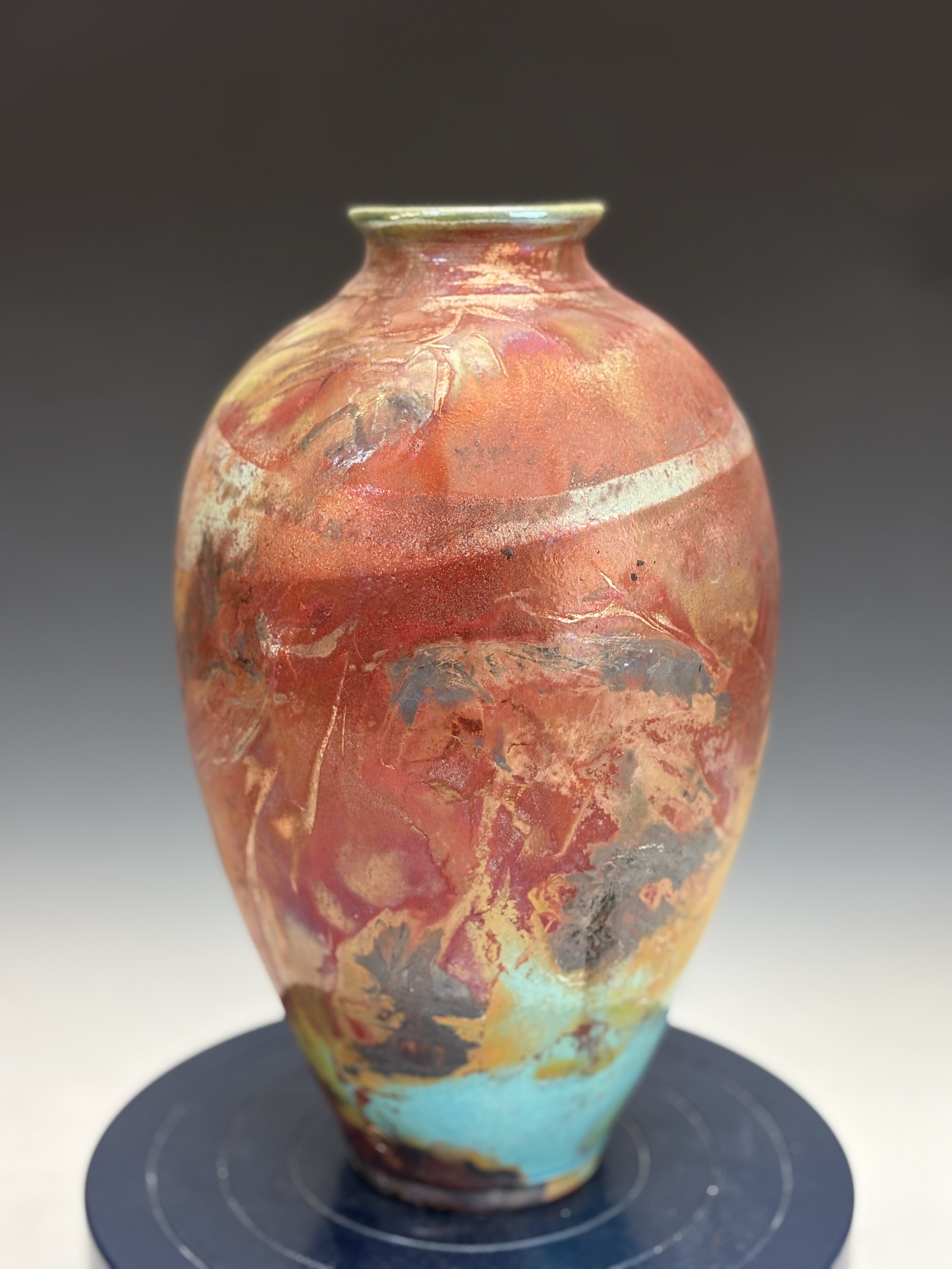 A colorful, glazed ceramic vase with an abstract, textured surface and a narrow neck, placed on a black circular stand against a plain background.
