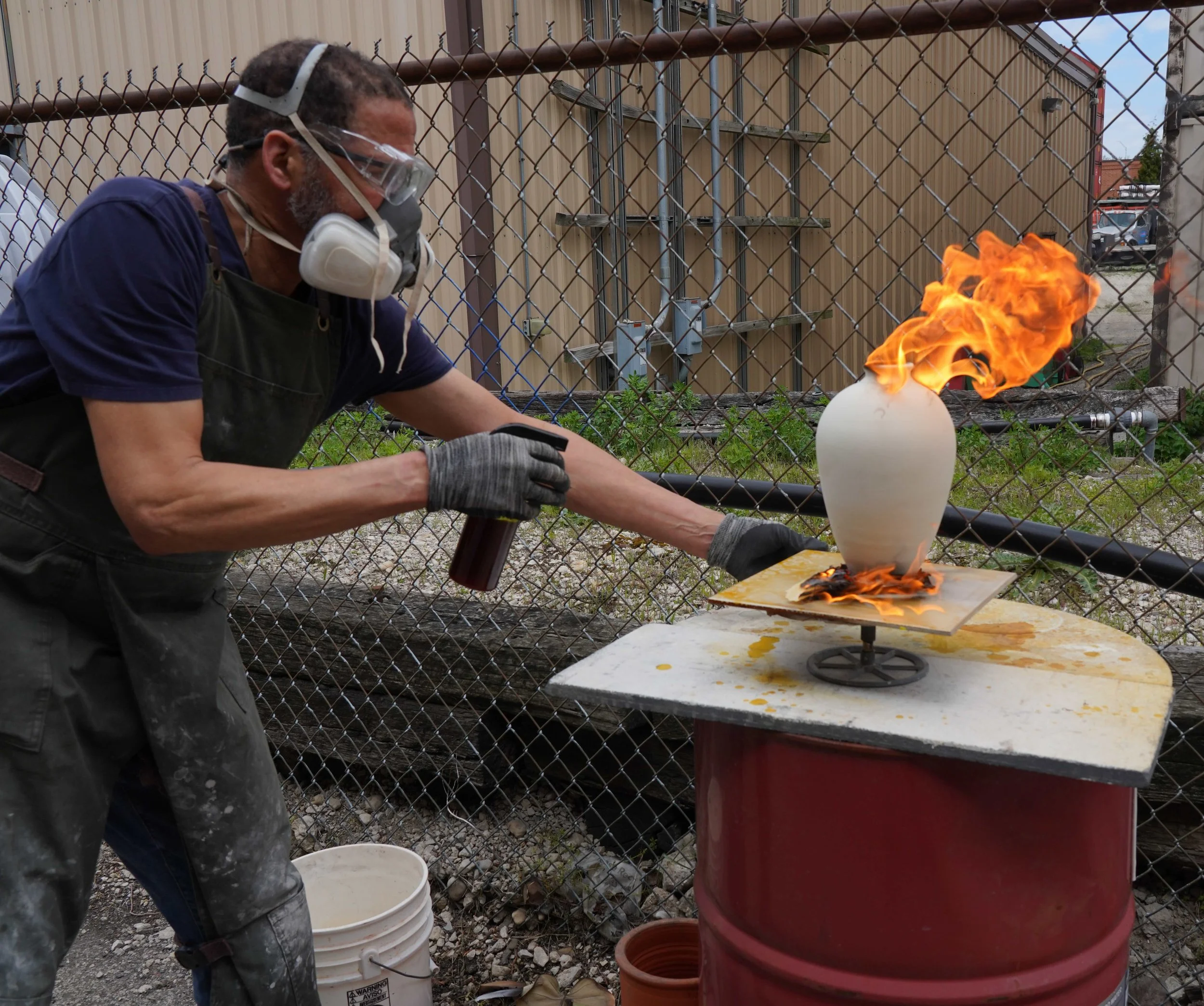 A man working on a chemistry experiment outdoors behind a chain-link fence, wearing safety goggles, a mask, and gloves, with flames coming from a ceramic vessel.