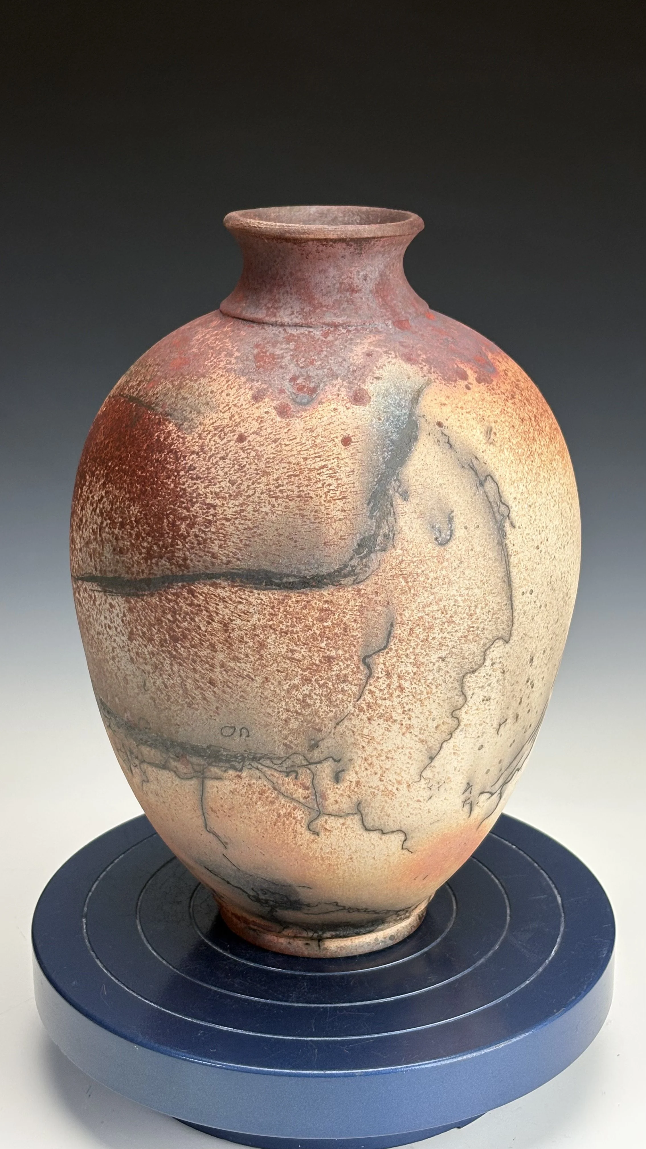 Ancient ceramic vase with a textured, reddish-brown surface and black and beige marbled patterns displayed on a black circular stand.