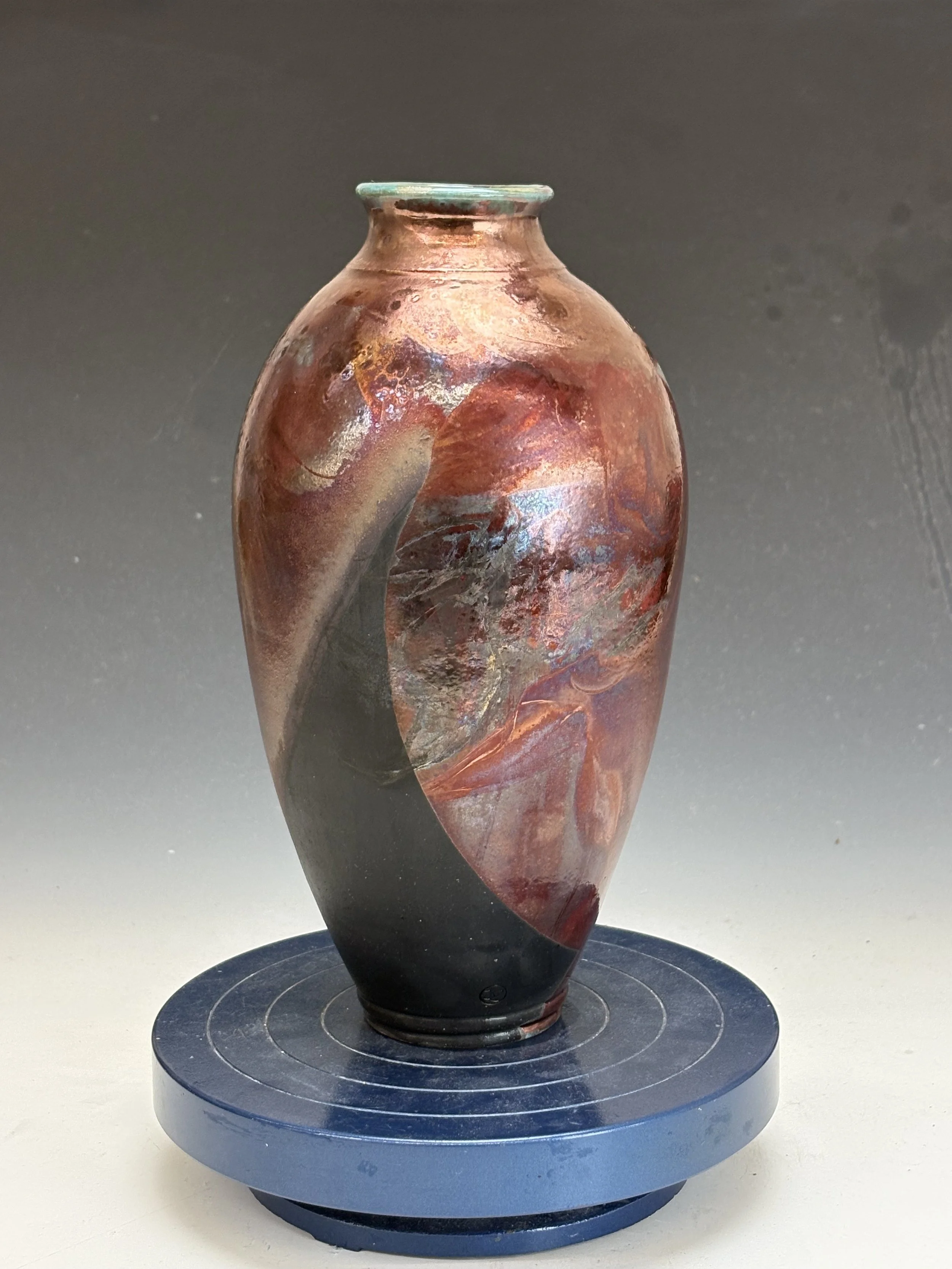 Ancient glass vase with curved body, reddish-brown and black swirls, displayed on a circular black stand against a plain background.