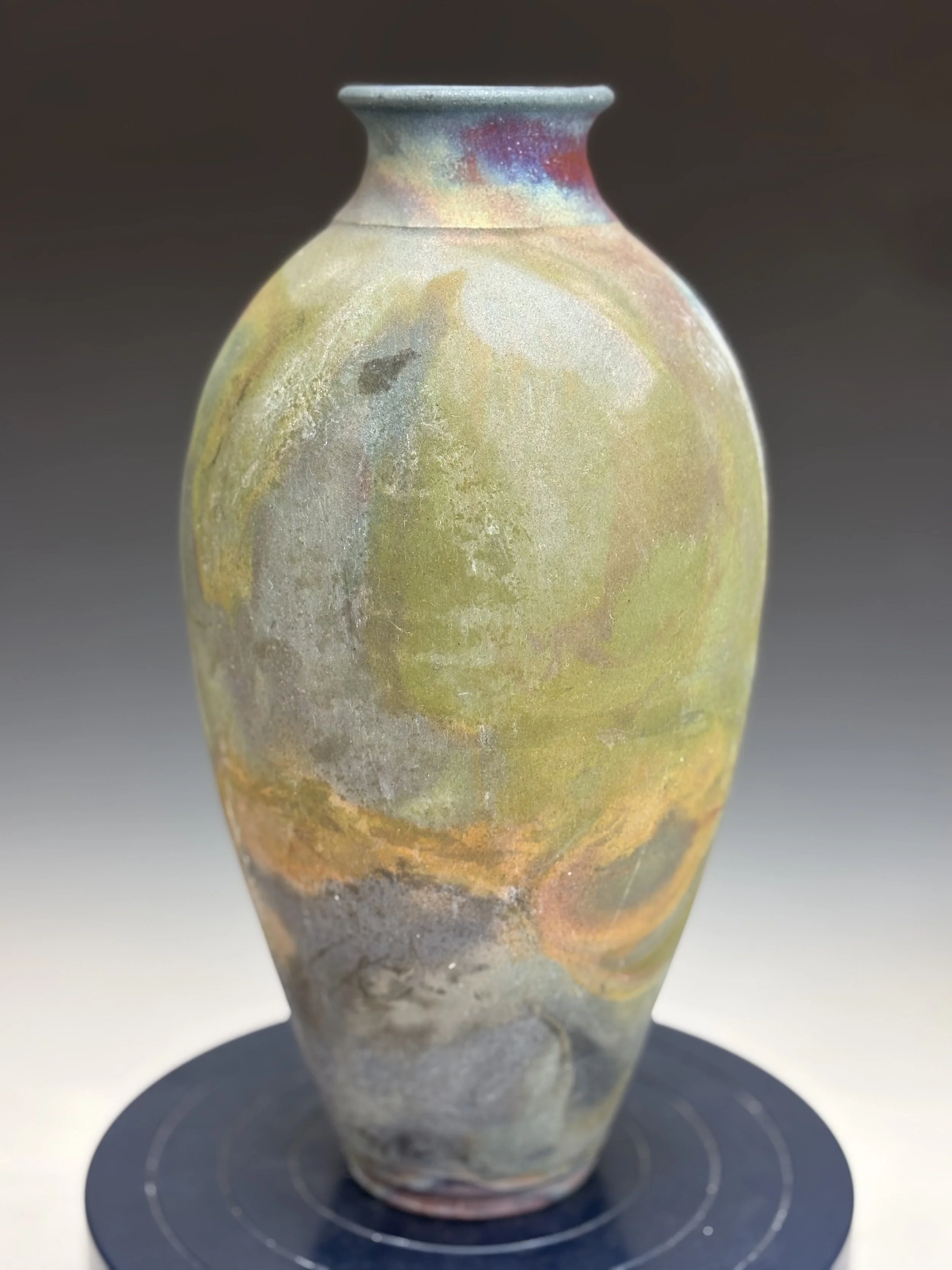 A ceramic vase with a rounded body and narrow neck, featuring a multicolored, iridescent glaze with shades of green, yellow, red, and blue, placed on a black circular stand.