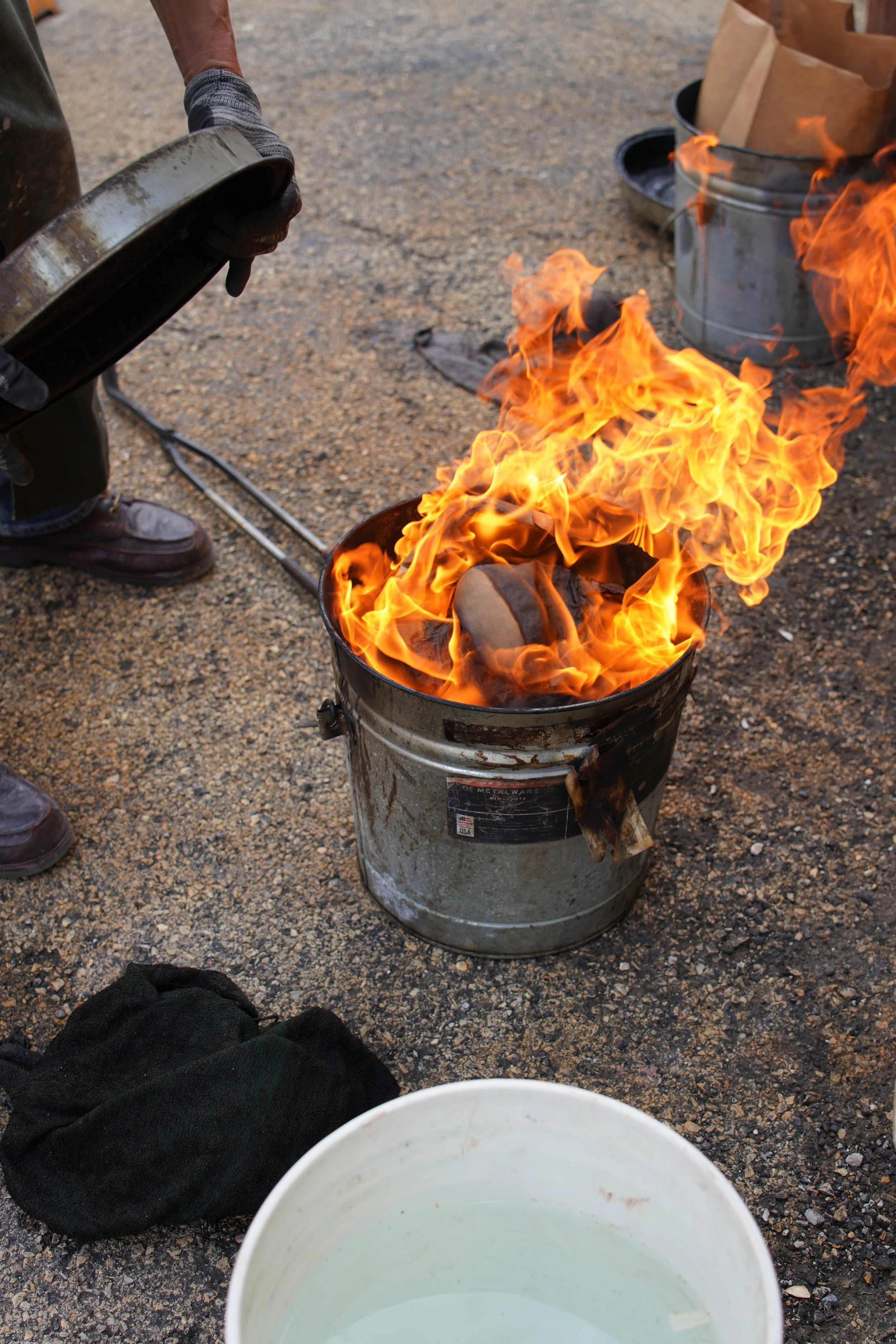 A person plunging a glowing-hot ceramic vase into a metal trash can that is on fire, creating bright orange flames. Various objects, including a paper bag and black containers, are nearby on a gravel surface.